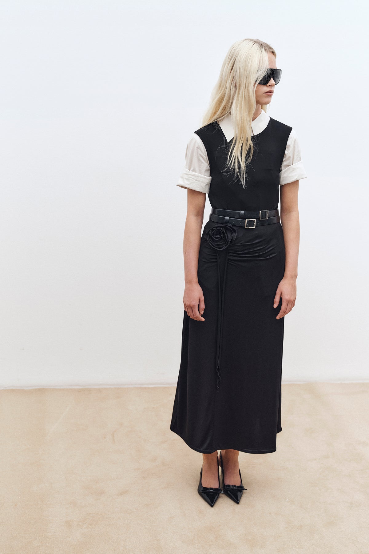Campaign portrait of a model in a black midi dress layered over a white collared blouse, with double belts, a rosette detail and oversized sunglasses.