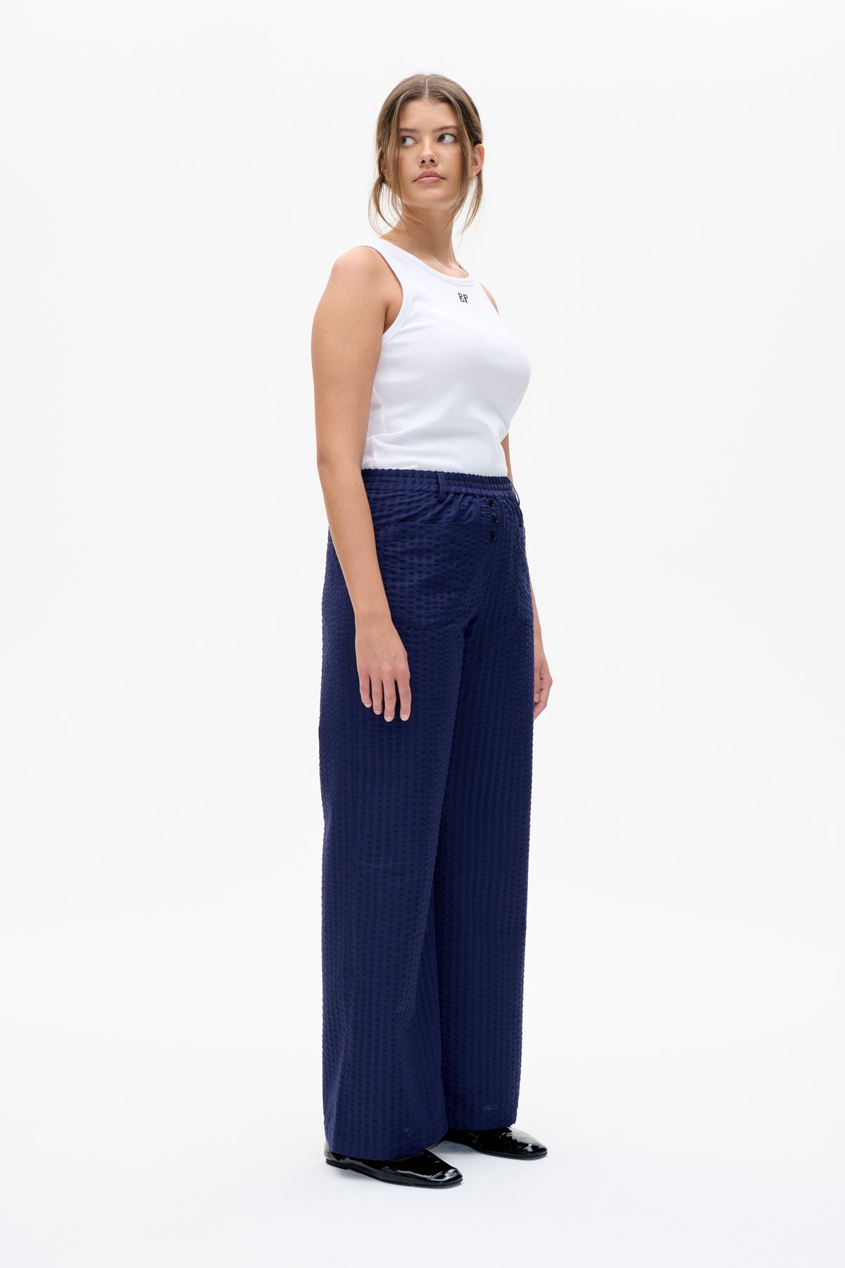 Model trägt ein weißes ärmelloses Top mit kleinem Brustemblem und eine strukturierte, marineblaue, weitgeschnittene Hose. Frontalansicht.
