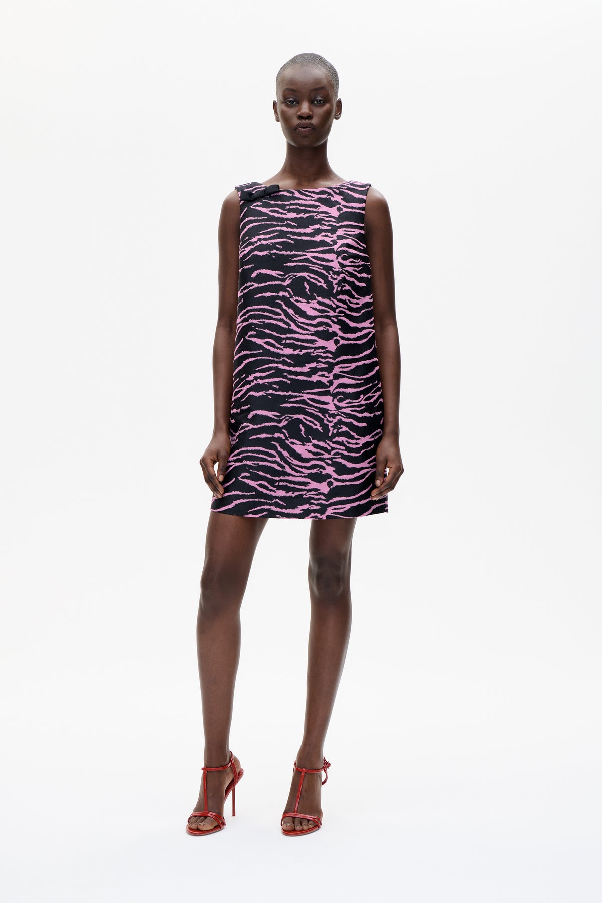 Front view of a sleeveless black mini dress with a pink wavy pattern and black shoulder bow, modeled against a white background.