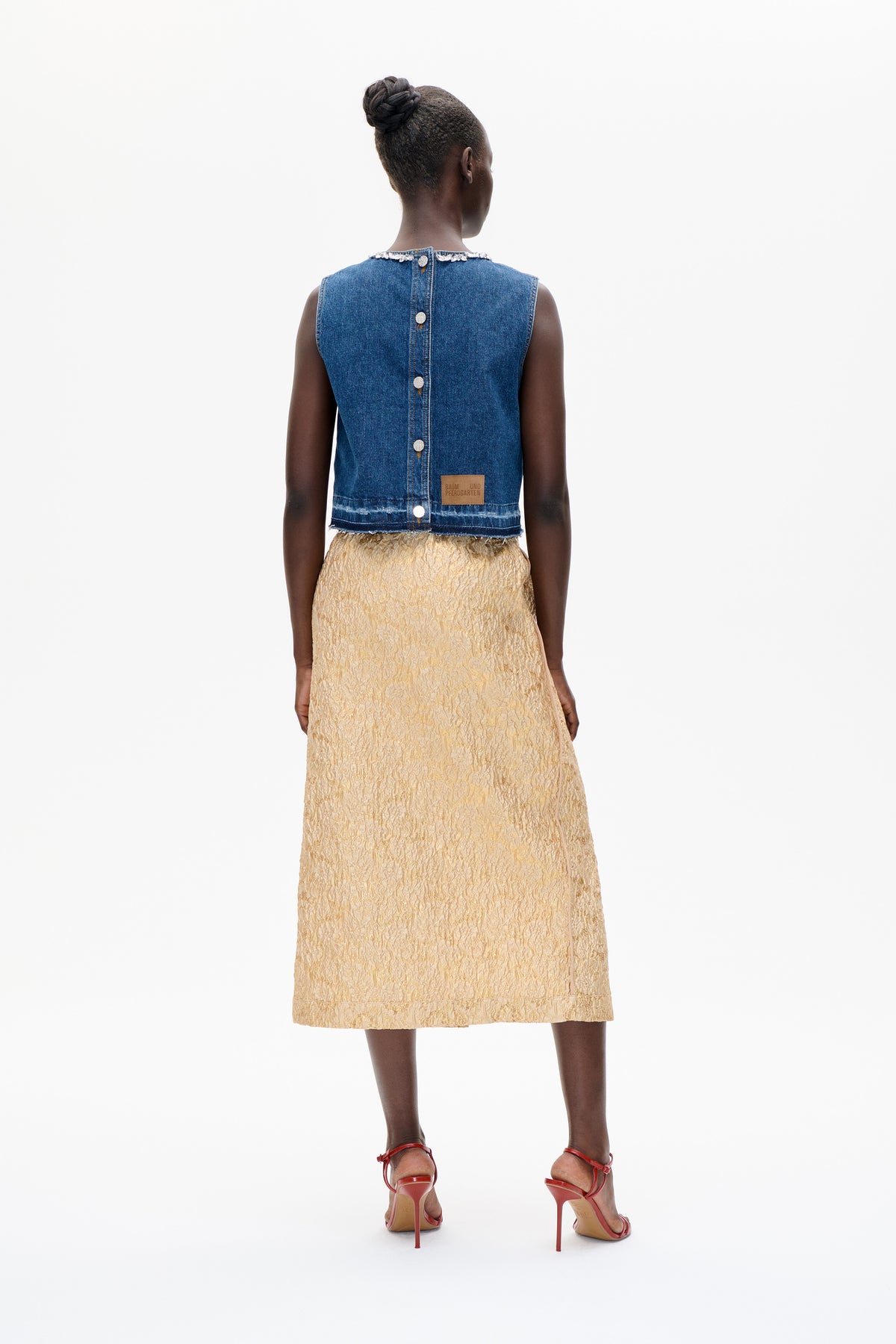 Achteraanzicht van een model dat een mouwloos denimvest met sierstenen langs de halslijn en knopen op de rug draagt, gecombineerd met een lichtgouden rok met structuur.