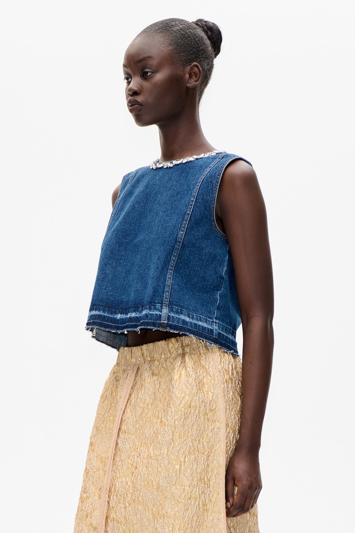 Model draagt een blauw mouwloos denim topje met onafgewerkte zoom en een halslijn met sierstenen, gecombineerd met een gestructureerde gouden rok.