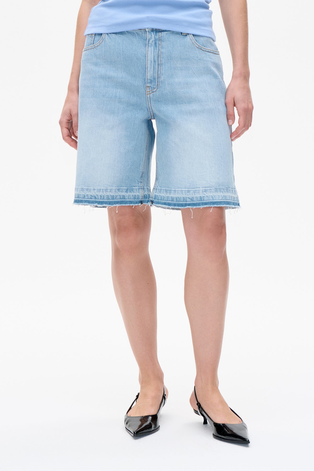 Front detail of light-wash denim shorts with wide legs, curved front pockets and frayed hems, styled with a pale blue top.