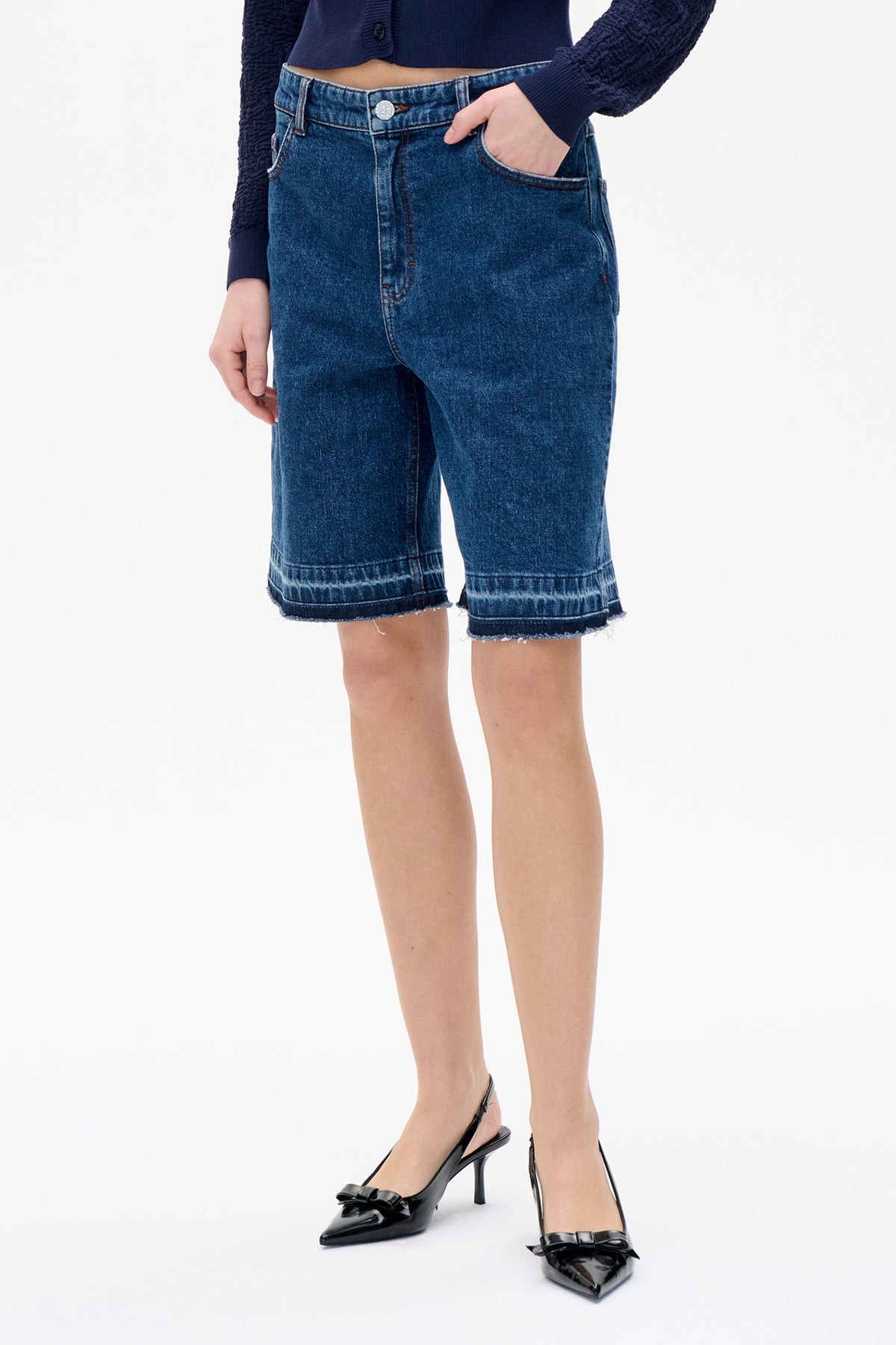 Front detail of dark blue denim Bermuda shorts with a button waist, front pockets and frayed hems, styled with a navy cardigan.