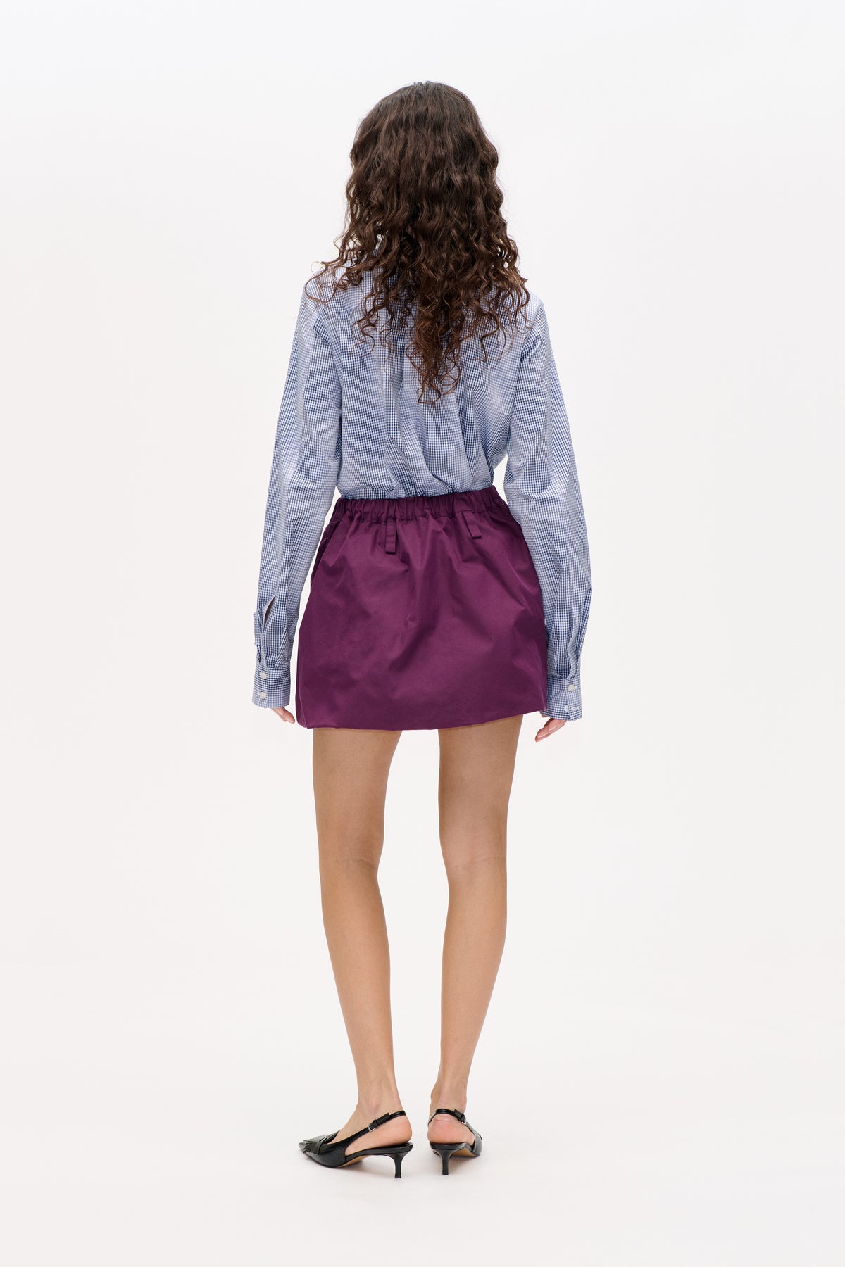 Back view of a model wearing a blue-and-white checked long-sleeve shirt and a plum mini skirt with an elastic waist.