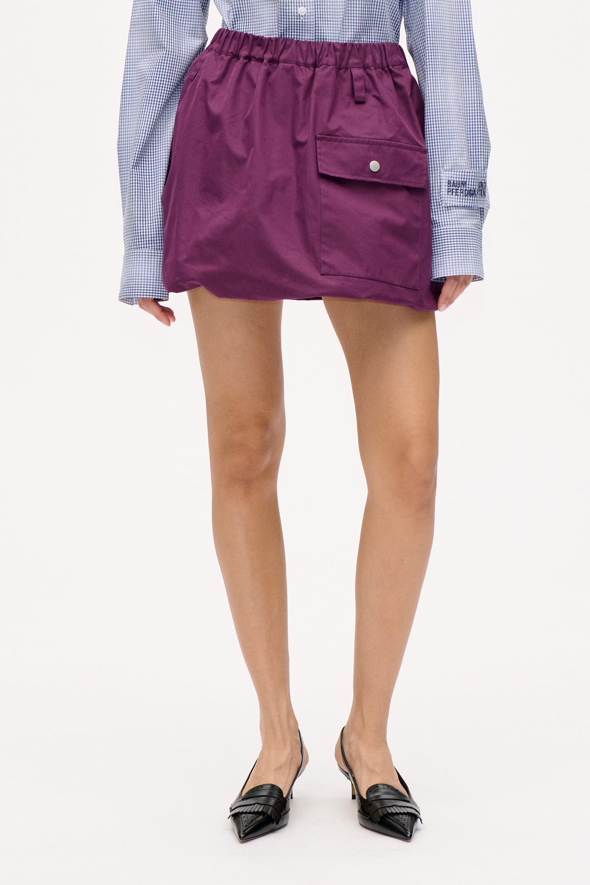 Close front detail of a plum mini skirt with an elasticated waist and oversized snap-fastened flap pocket, worn with a blue checked shirt.