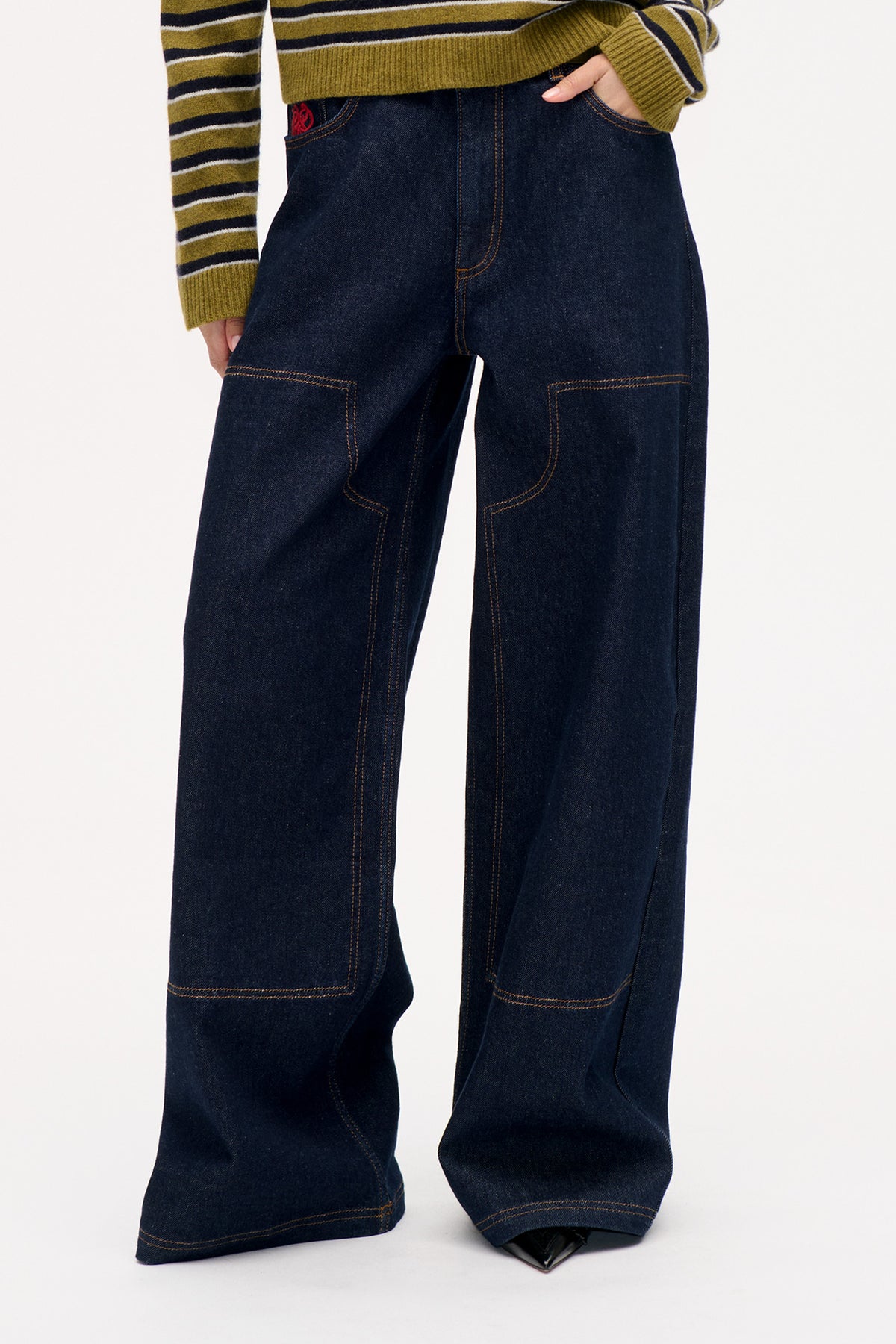 Front detail of dark indigo wide-leg jeans with contrast stitching and large rectangular patch pockets, styled with an olive striped sweater.