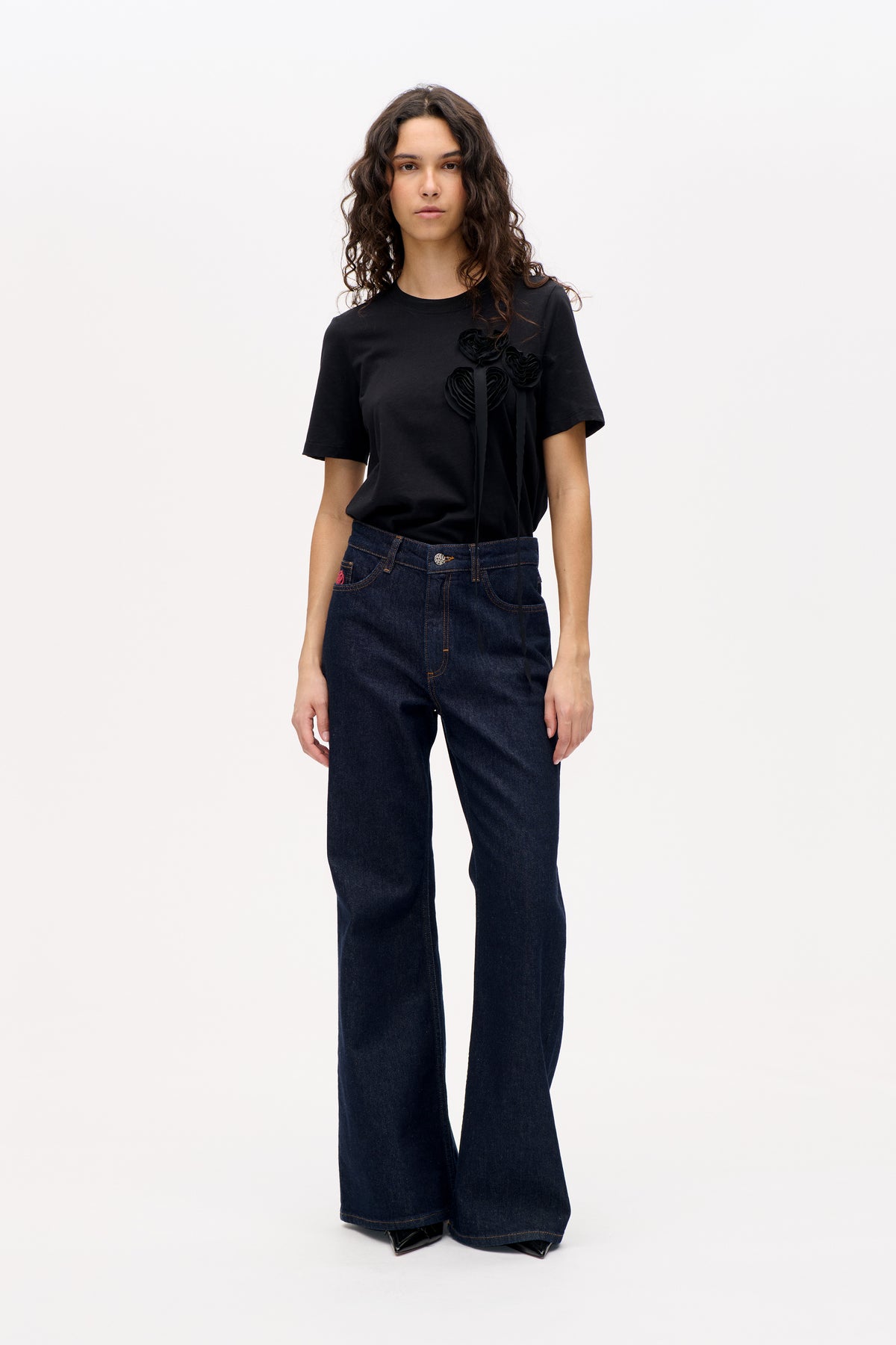 Vorderansicht eines Models in einem schwarzen, kurzärmeligen T-Shirt mit mehrlagigen Stoffblüten-Applikationen und dunklen Jeans mit weitem Bein.
