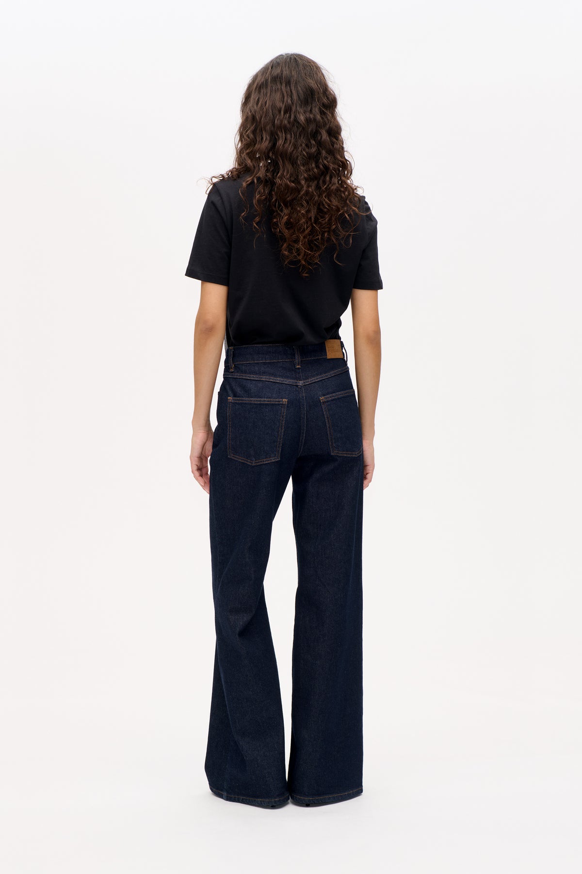 Back view of a model wearing a plain black short-sleeve T-shirt and dark indigo wide-leg jeans with contrast stitching and back pockets.