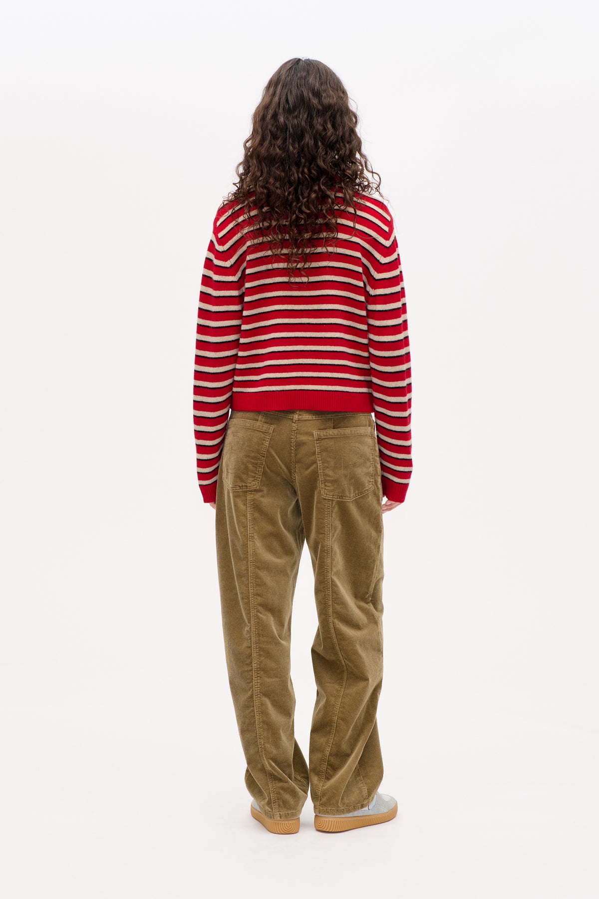 Back view of a model wearing a red sweater with cream and navy horizontal stripes and loose brown corduroy trousers.