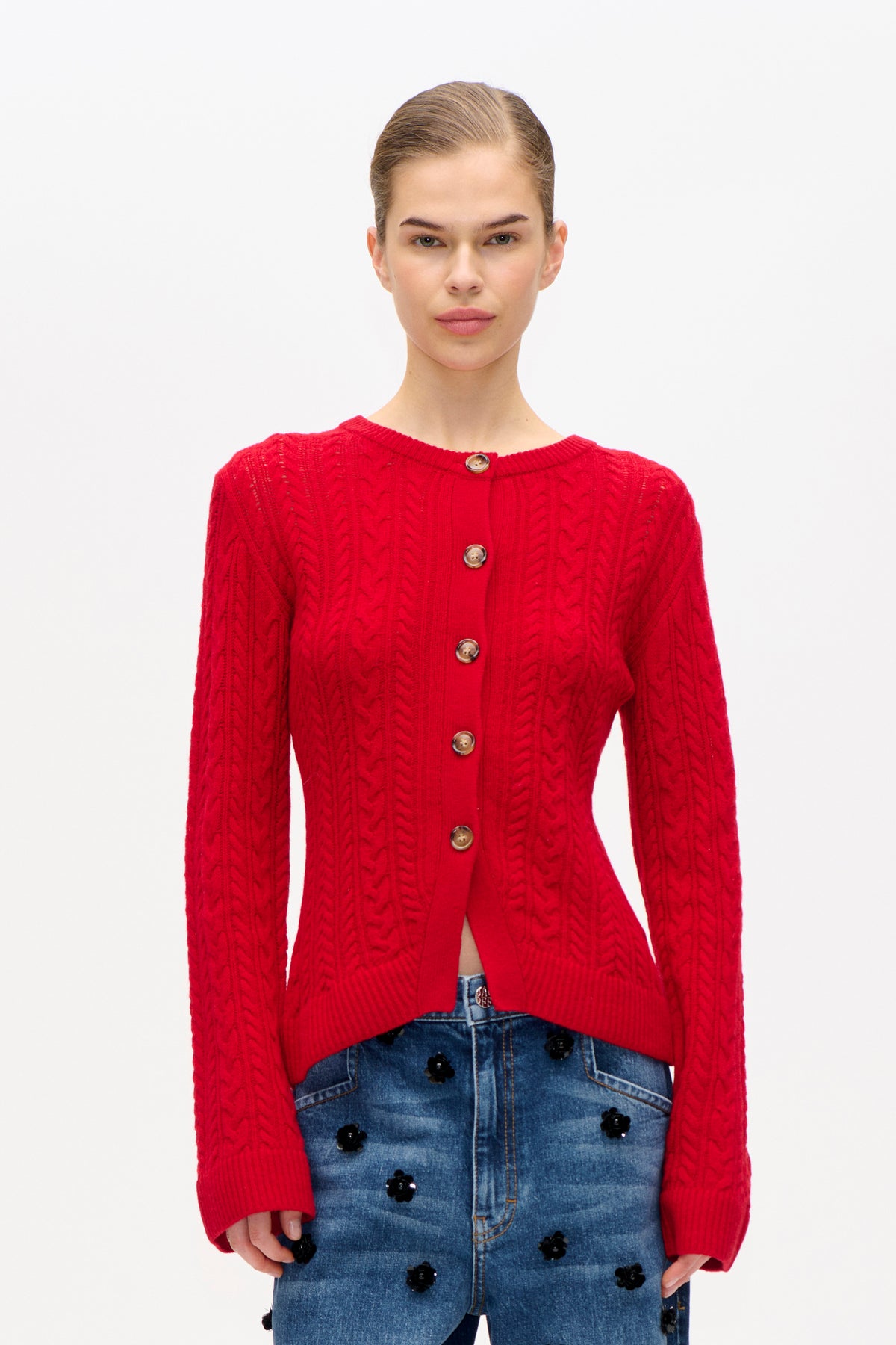 Front view of a model wearing a bright red cable-knit cardigan with brown buttons and a blue denim skirt decorated with black flowers
