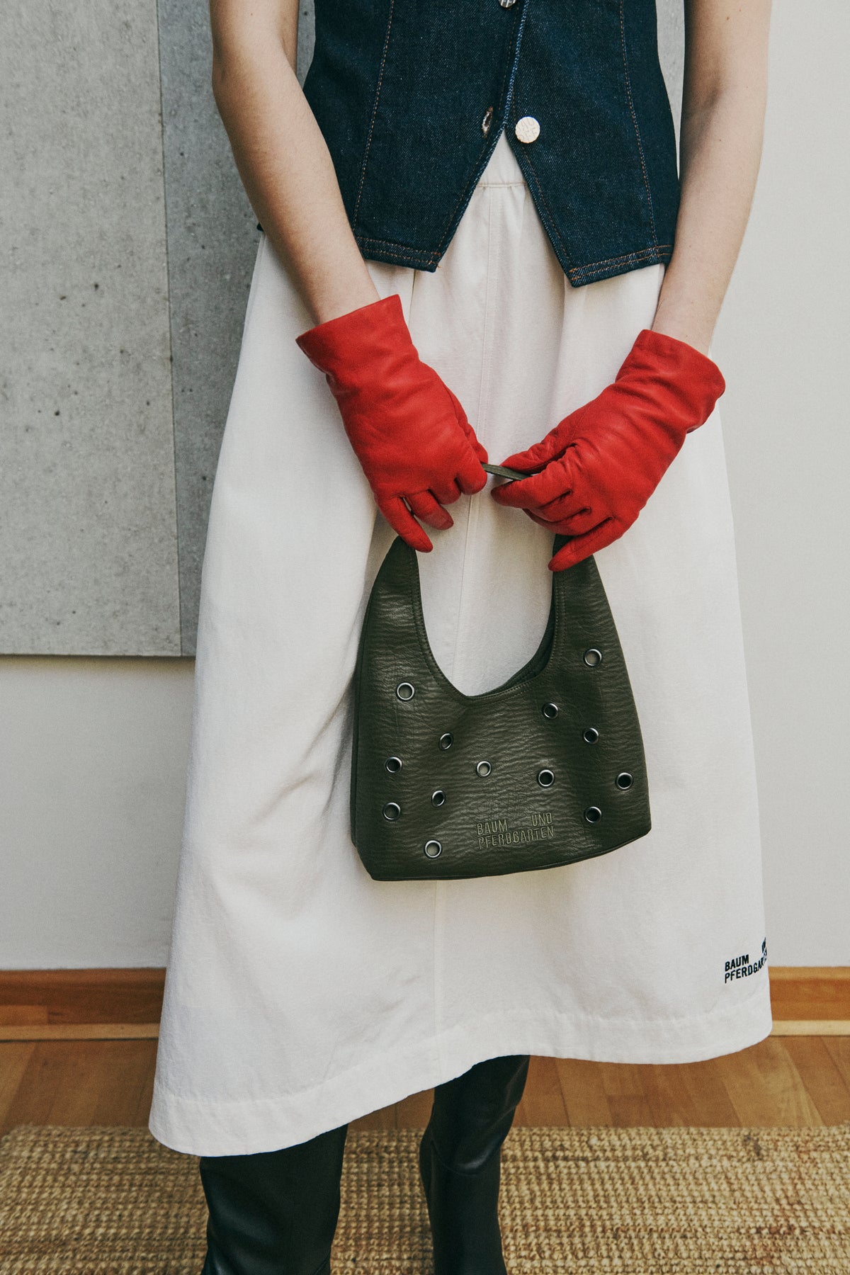 Cropped campaign view of a model holding a dark green eyelet-studded bag, styled with red gloves, a white apron and dark denim vest.