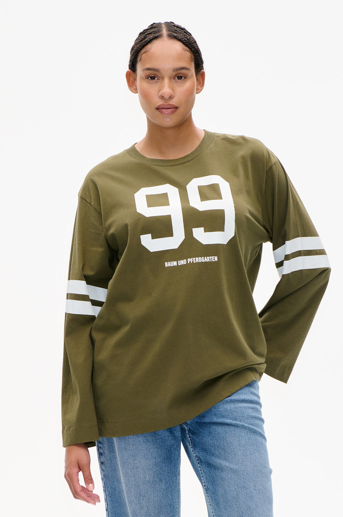 Vue de face d'un t-shirt vert olive à manches longues, avec un grand motif blanc "99", le lettrage Baum und Pferdgarten et des bandes blanches aux manches, porté avec un jean bleu.