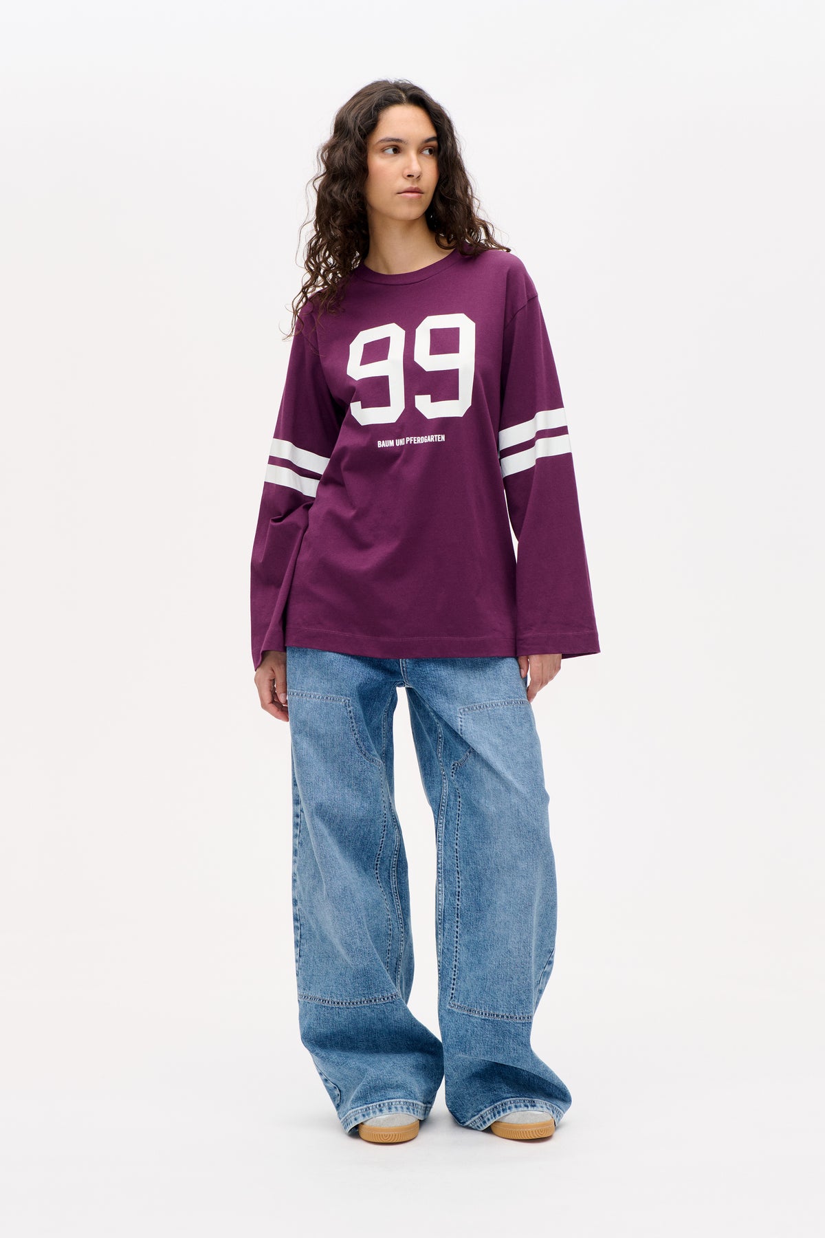 Front view of a model wearing a plum long-sleeved sweatshirt with a large white 99 graphic and white sleeve stripes, paired with loose blue jeans.