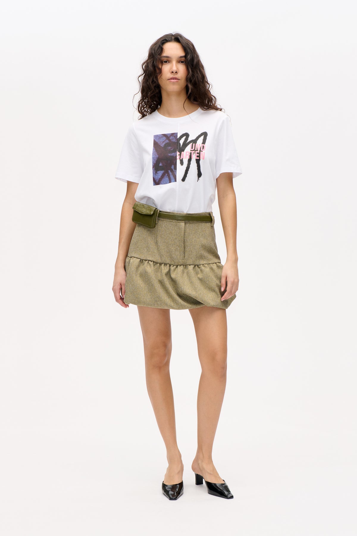 Model trägt ein weißes kurzärmeliges T‑Shirt mit auffälligem Print in schwarz, blau und pink, einen khakifarbenen Tweed-Minirock und eine olivgrüne Bauchtasche.