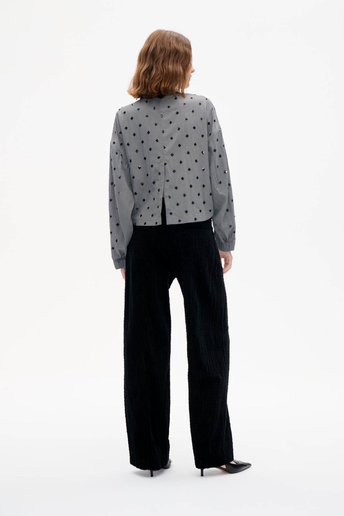 Rückenansicht eines Models, das eine graue, langärmelige Bluse mit vereinzelten schwarzen Verzierungen und eine schwarze, weit geschnittene Hose trägt.