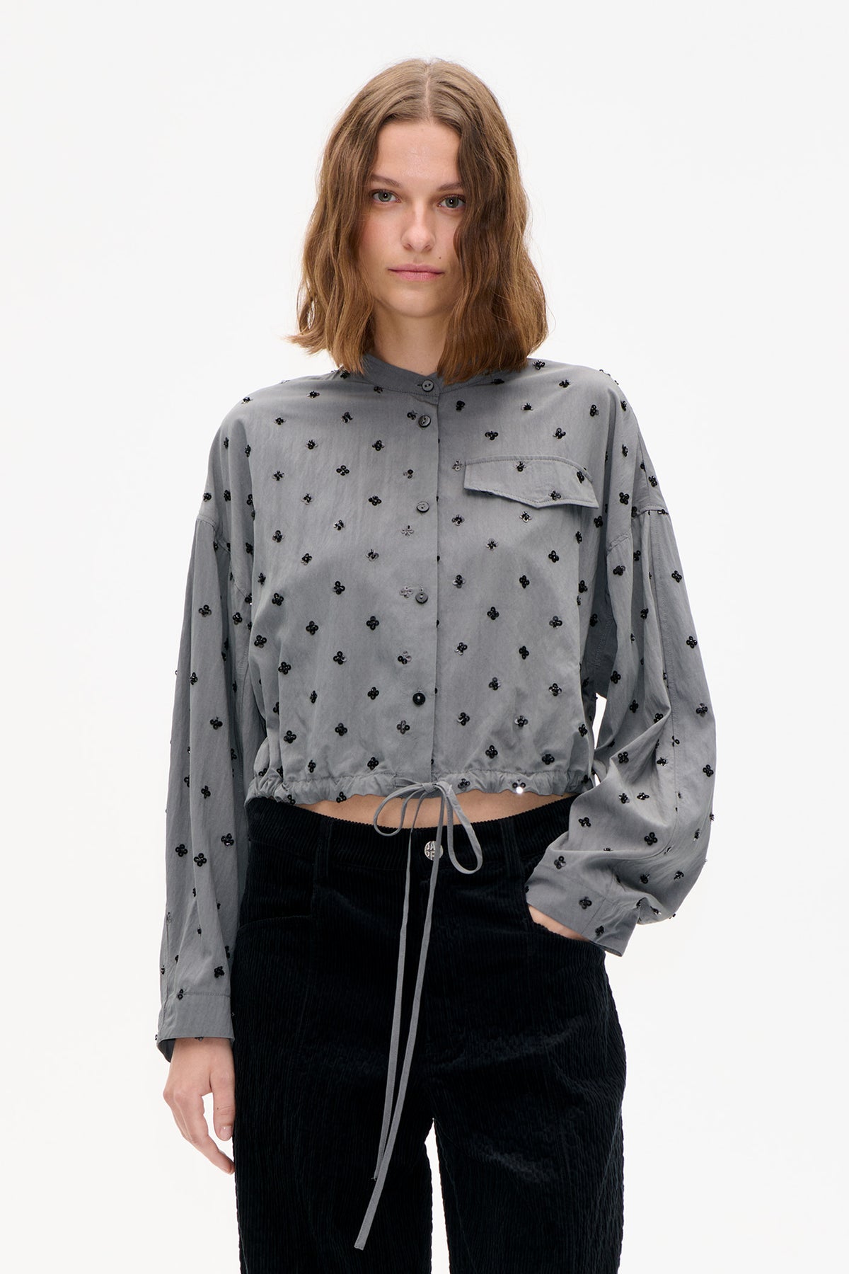 Front view of a cropped grey button-front shirt with scattered black floral embellishments, a chest flap pocket and gathered drawstring hem.