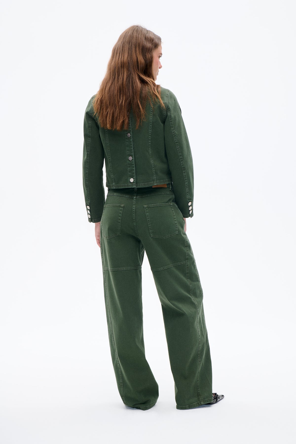 A model wears a dark green denim jacket with silver buttons and matching wide-leg trousers, shown from behind.