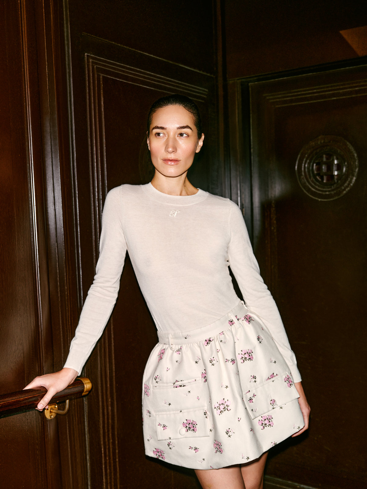 Model wears a white long-sleeved top and an ivory mini skirt with scattered pink floral print and large patch pockets.