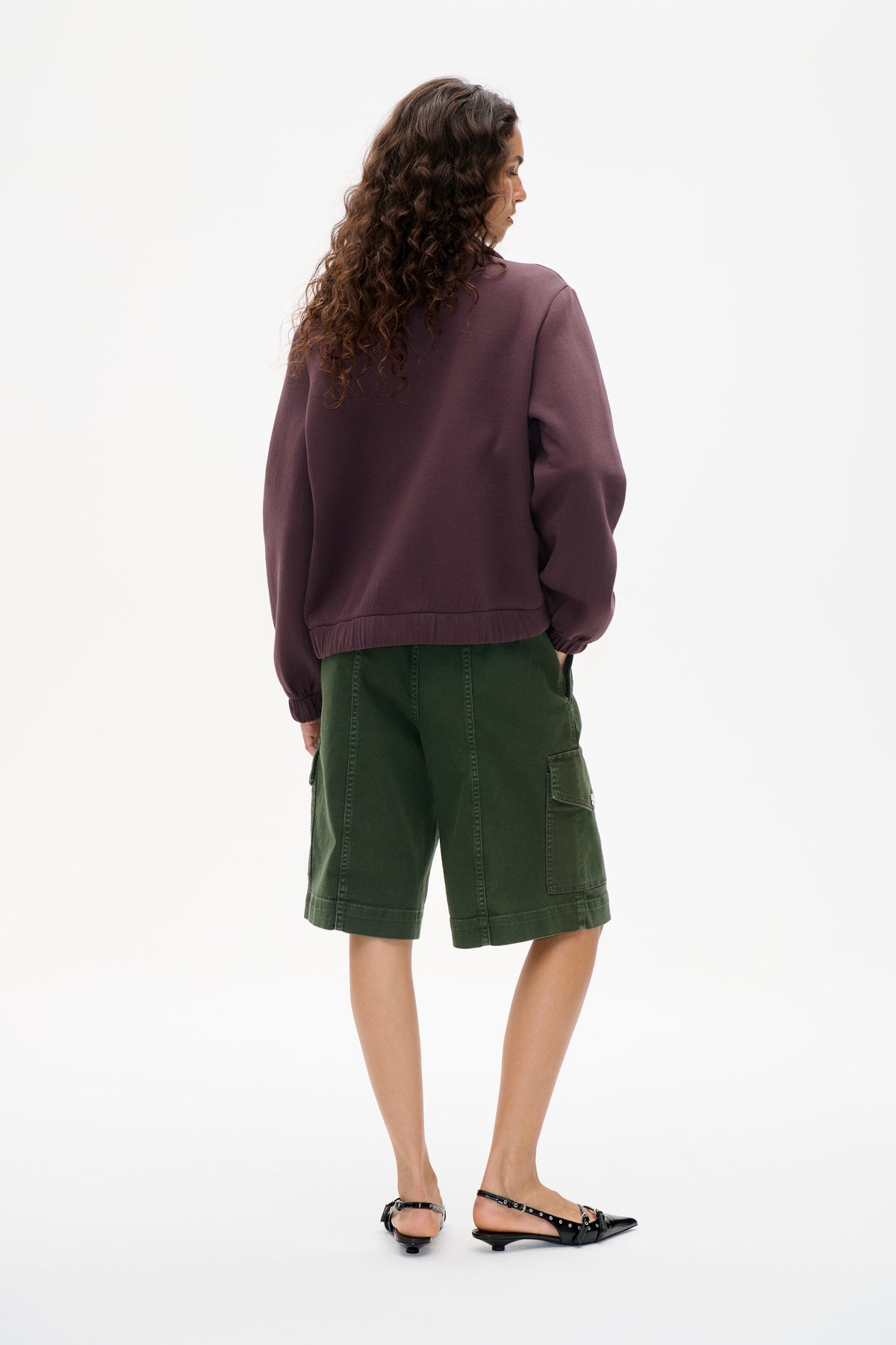 Back view of a model wearing a muted plum sweatshirt with gathered cuffs and hem, paired with dark green knee-length cargo shorts.