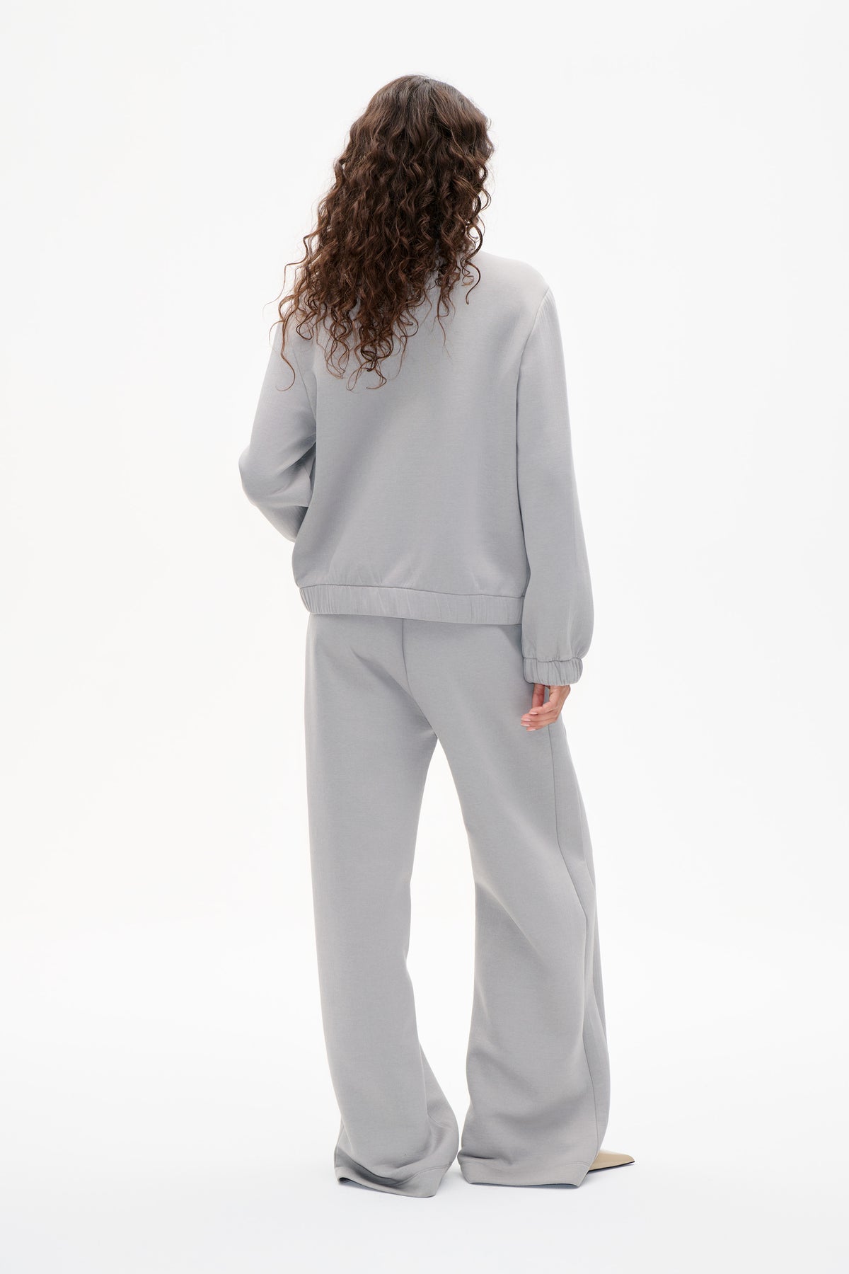 Back view of a model wearing a light grey sweatshirt with elastic cuffs and hem, paired with matching wide-leg trousers.
