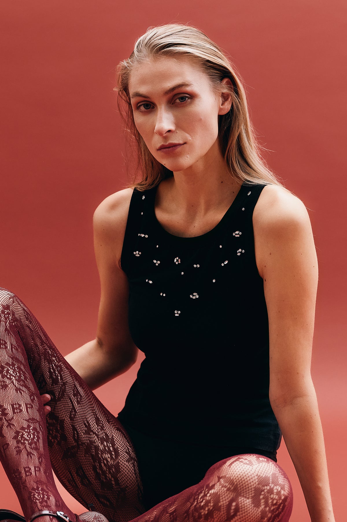 Campaign portrait of a seated model wearing a black sleeveless top with scattered sparkling embellishments and burgundy patterned sheer tights against a coral backdrop.