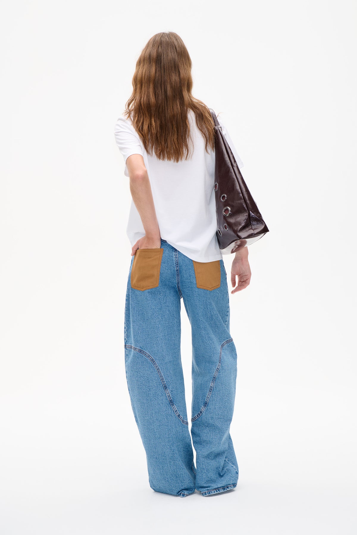 Back view of a model wearing a white short-sleeve T-shirt and wide-leg blue jeans with tan patch pockets and curved seams, carrying a dark brown grommet tote.
