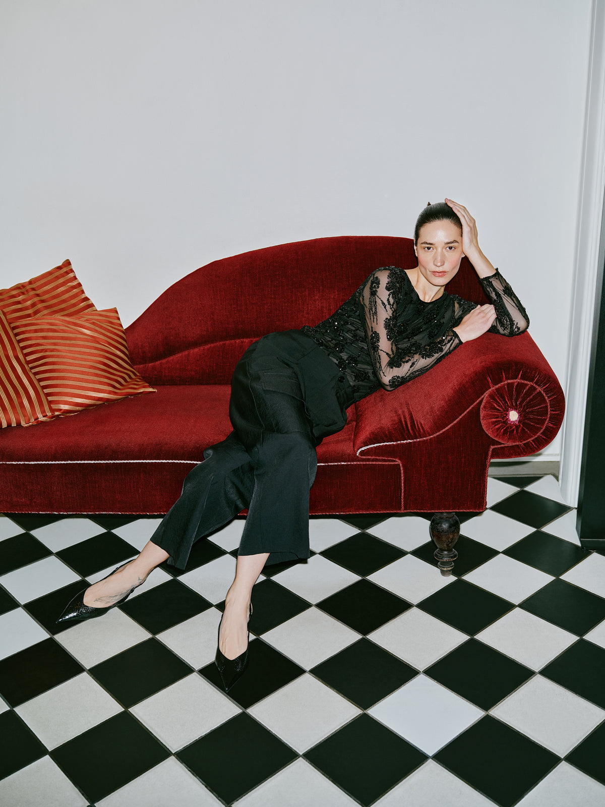 Model reclining on a red velvet sofa in a sheer black floral lace long-sleeve top, black trousers and pointed shoes.