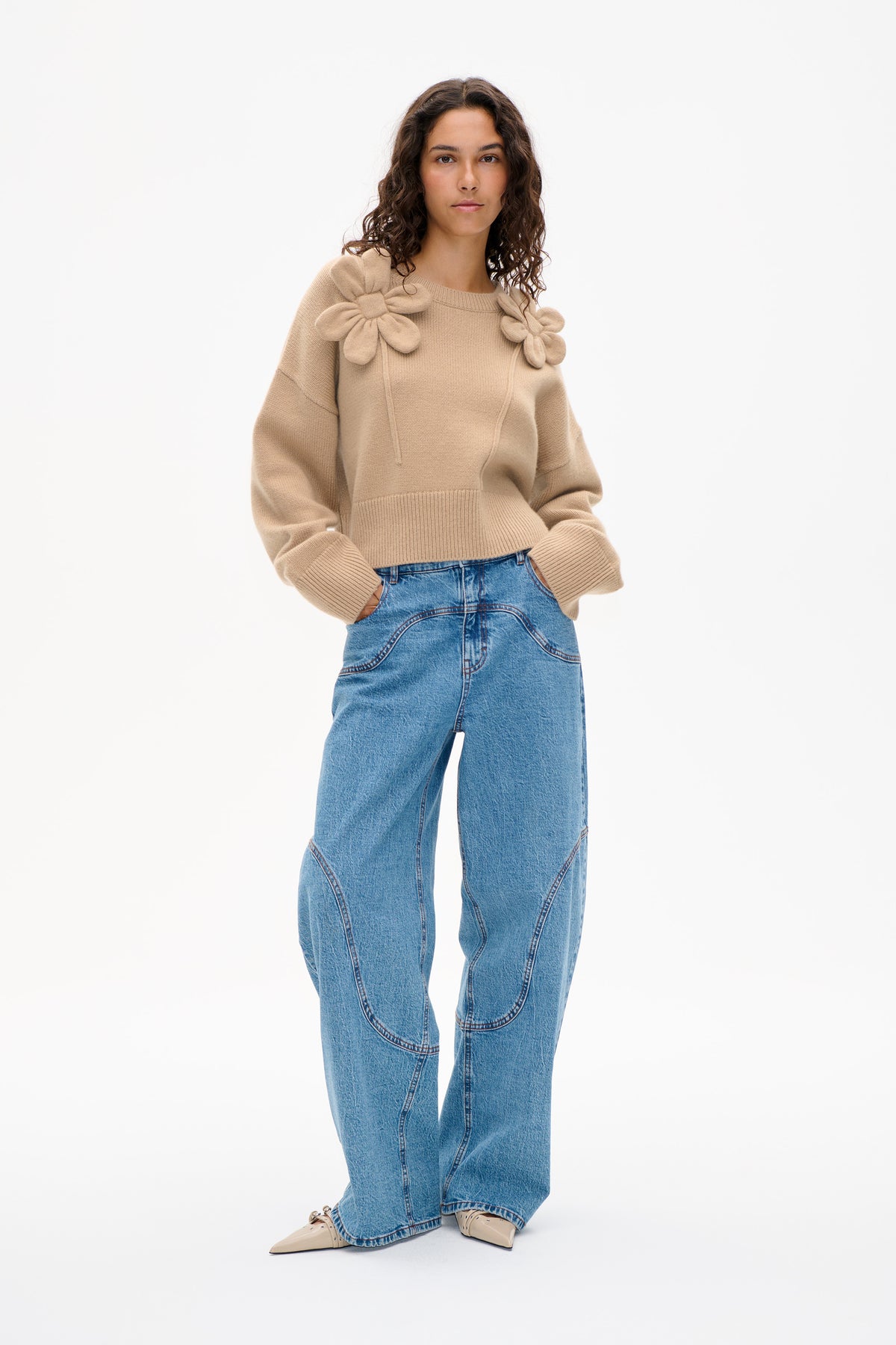 Vorderansicht eines beigen Strickpullovers mit übergroßen Blumenapplikationen an den Schultern, kombiniert mit weiten blauen Jeans.