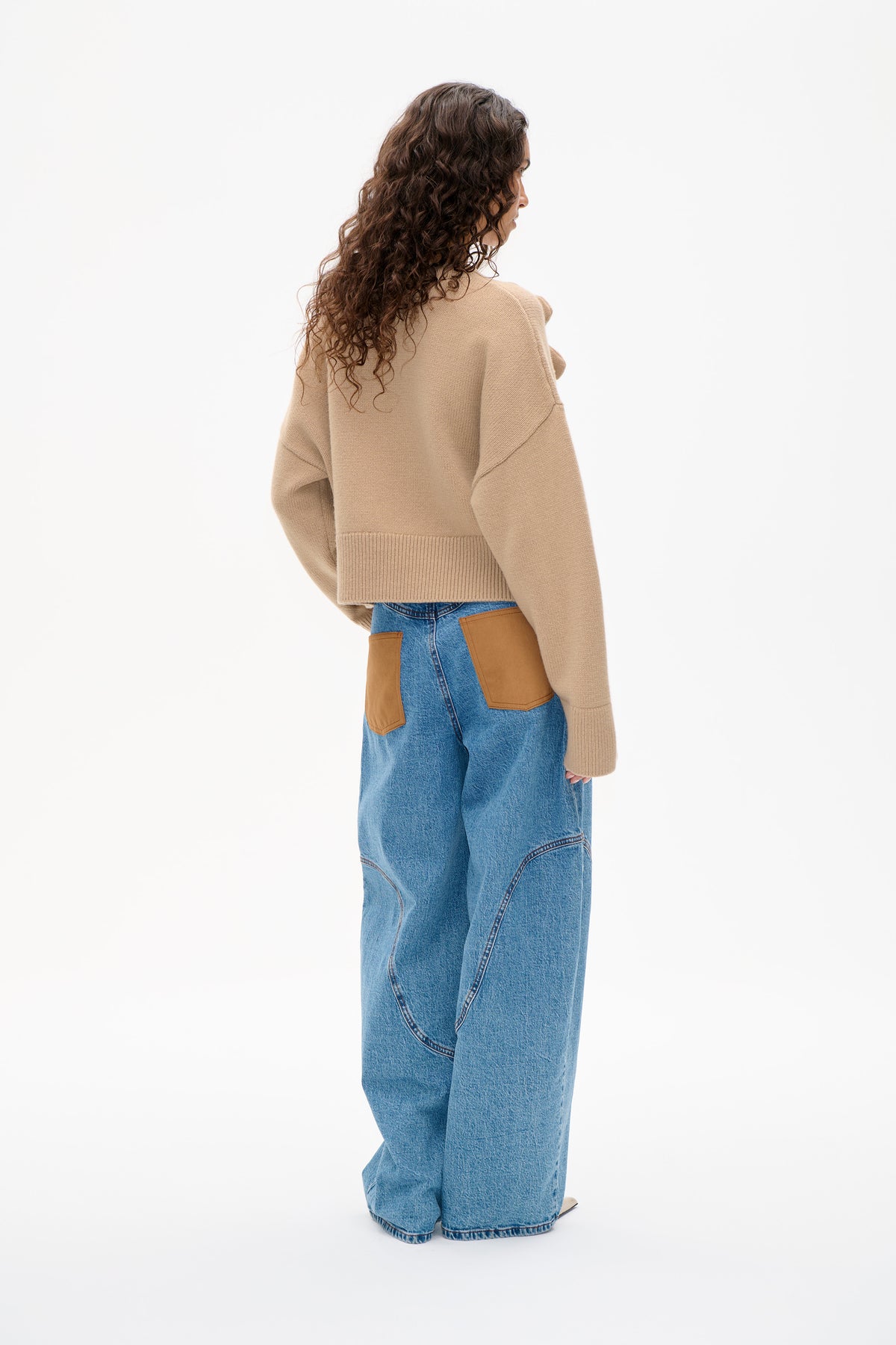 Model zeigt den Rücken eines beigefarbenen Strickpullovers mit überschnittenen Schultern und geripptem Saum, kombiniert mit blauen Jeans mit hellbraunen aufgesetzten Taschen.