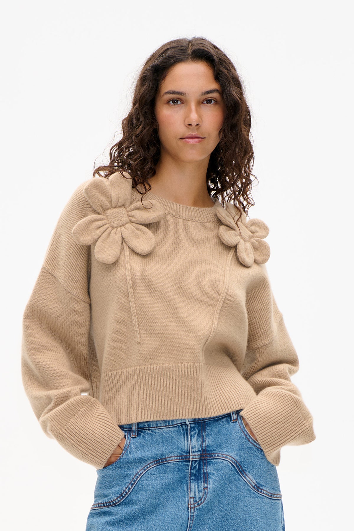 Front view of a model wearing a beige rib-knit sweater with oversized knitted flower appliqués around the neckline and blue jeans.
