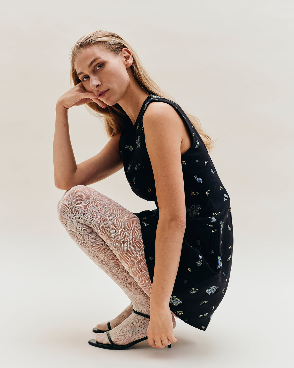 Campagneportret van een zittend model in een zwarte mouwloze jurk met kleine blauwe en crèmekleurige bloemen, lichte opengebreide panty's en zwarte hakken.