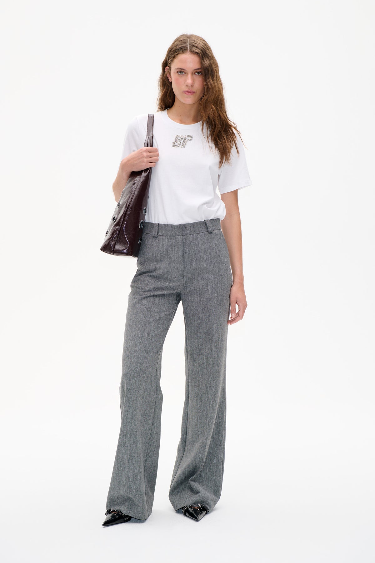 Model trägt ein weißes, kurzärmeliges T-Shirt mit verzierten Initialen, dazu graue, weit geschnittene Hosen und eine glänzende, dunkle Schultertasche.