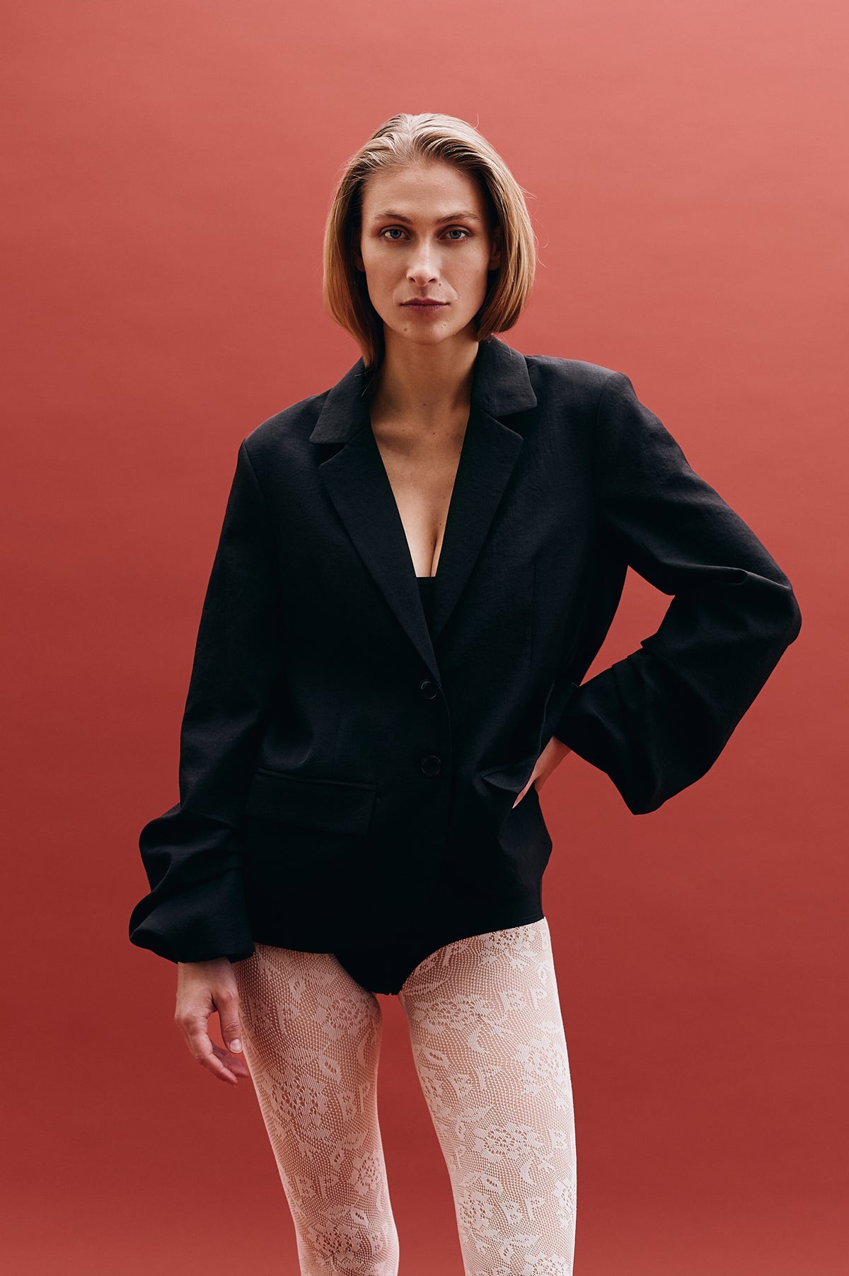 Model posed against a coral backdrop in a black button-front blazer and pale cream lace trousers.