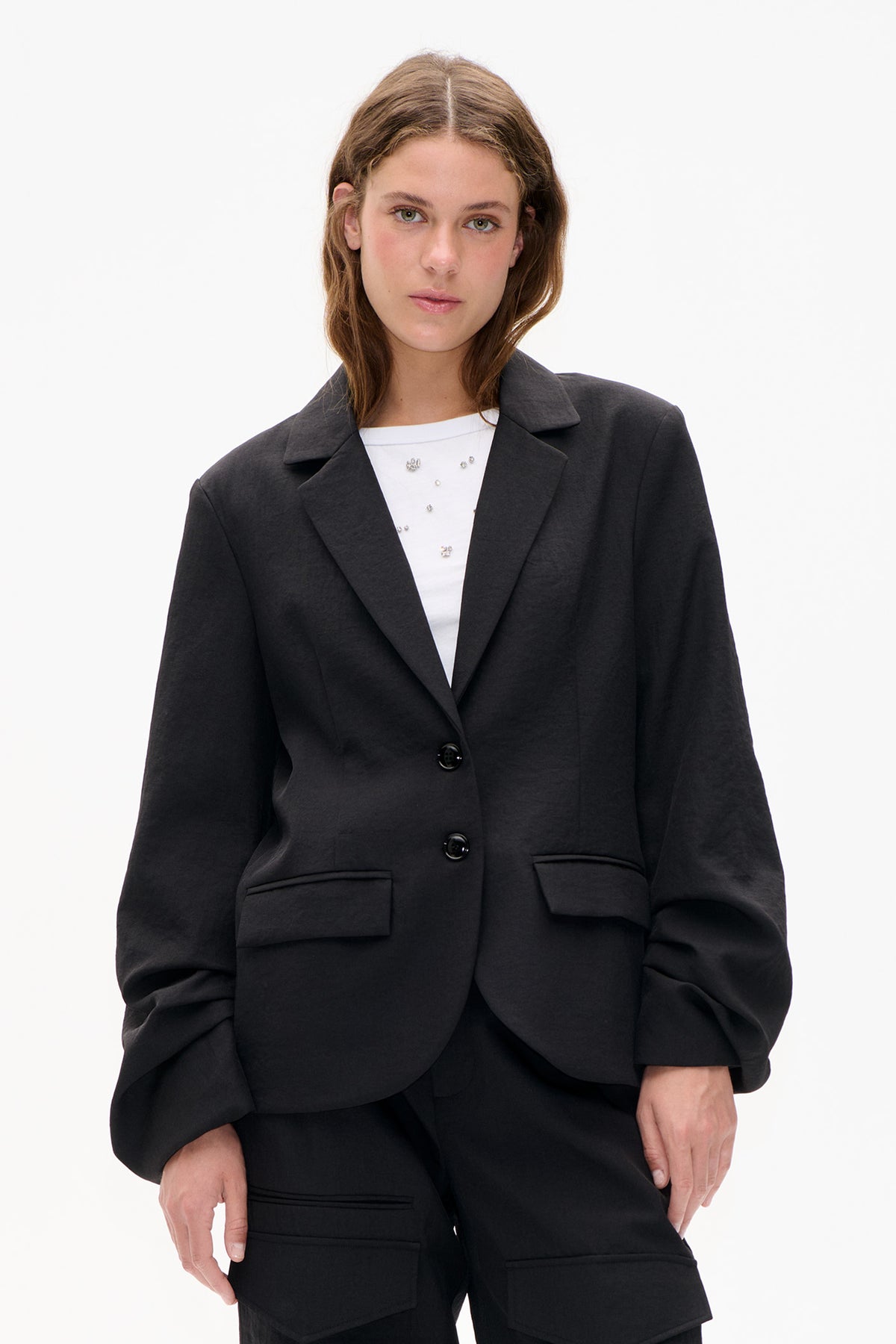 Front view of a model wearing a charcoal blazer with wide lapels, two buttons and flap pockets over a white embellished top.