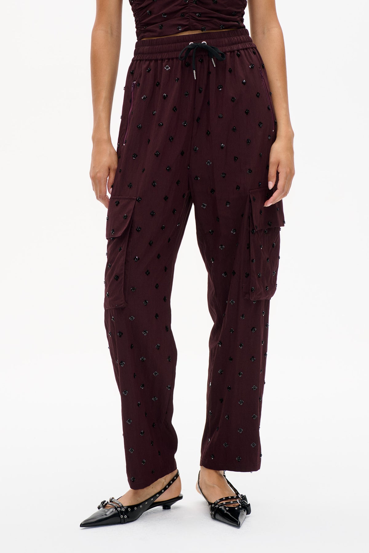 Detail van de voorkant van een bordeauxrode broek met trekkoord, verspreide zwarte bloemdetails, cargozakken en taps toelopende pijpen.