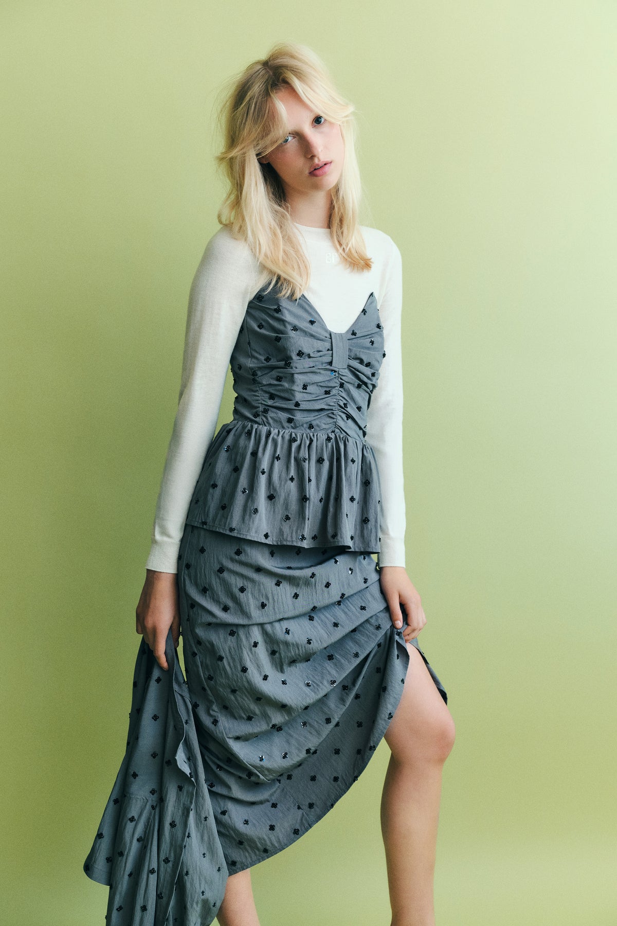 Model trägt ein graues Kleid mit gerafftem Oberteil und schwarzen Blumenapplikationen über einem weißen Langarmtop vor hellgrünem Hintergrund.