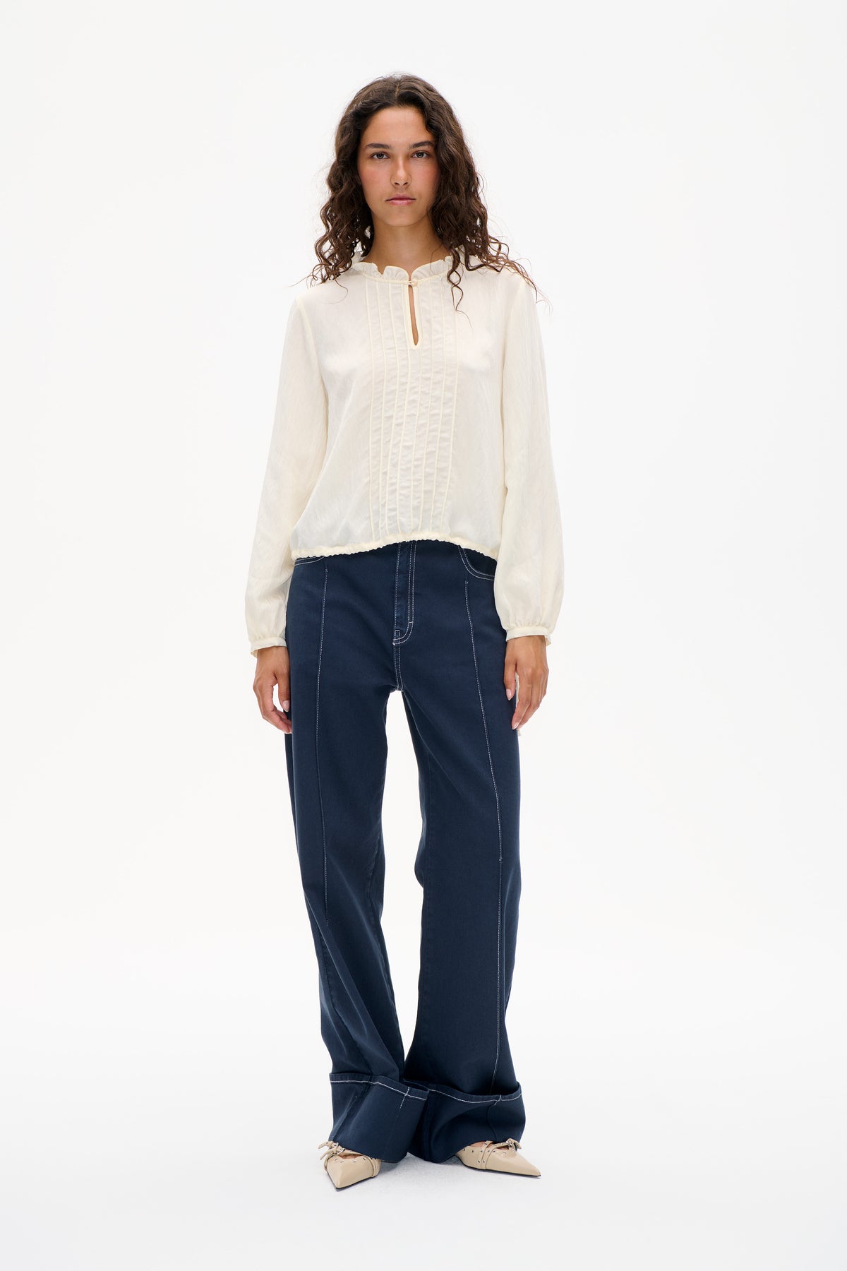 Front view of a model wearing an ivory long-sleeved blouse with a ruffled neckline, pintuck panels and a small keyhole opening, paired with dark blue jeans.