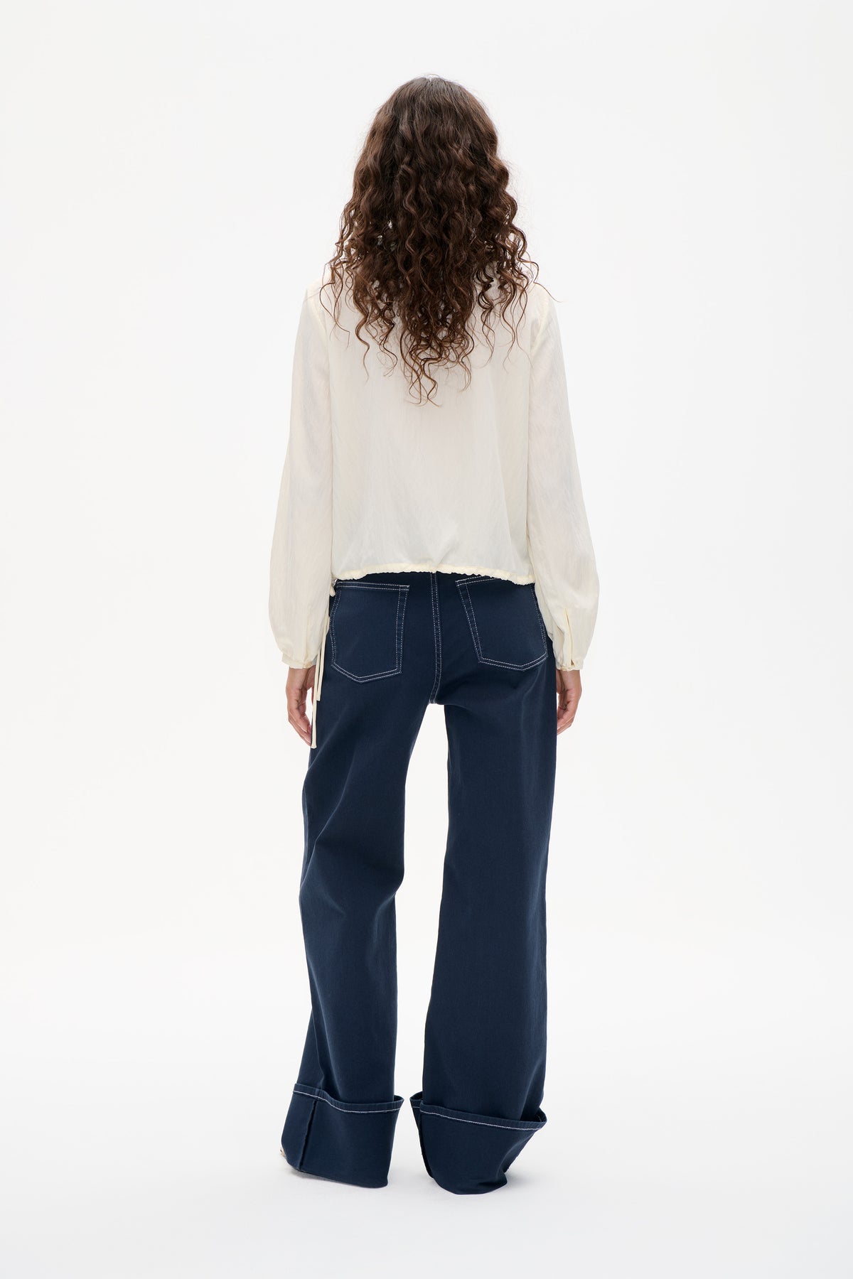 Back view of an ivory long-sleeved blouse with dark navy wide-leg trousers, oversized turned-up hems, back pockets and contrast stitching.