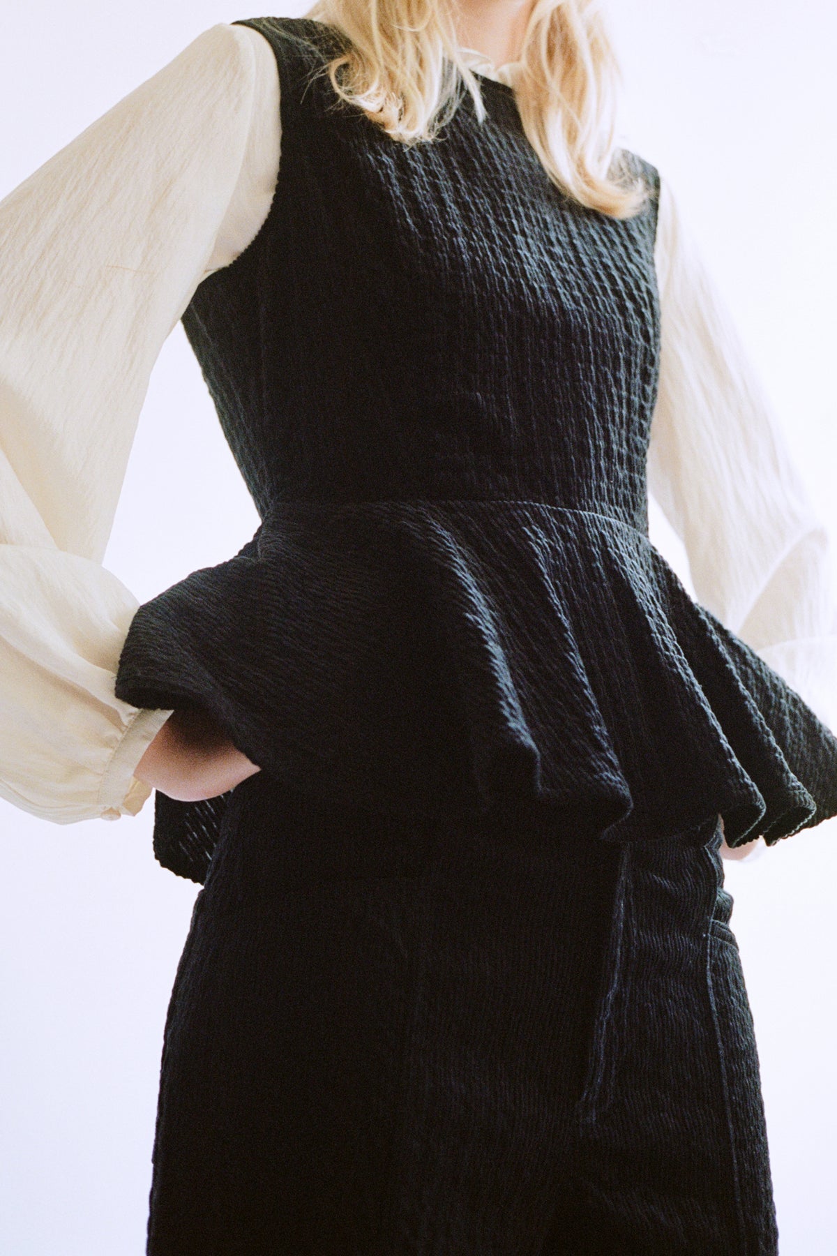 Close-up campaign portrait of a model in a black ribbed sleeveless dress layered over a cream long-sleeved blouse.