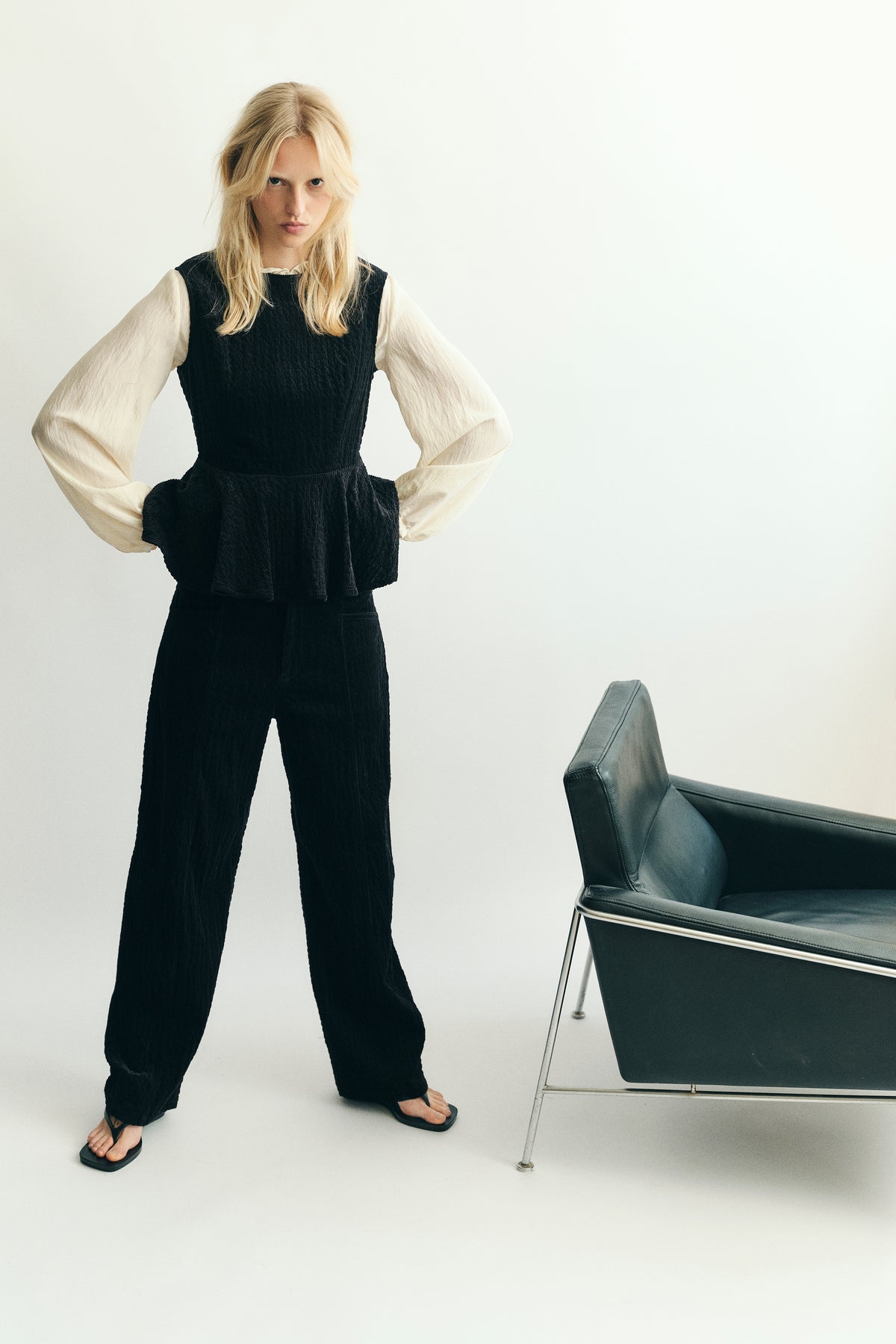 Model wearing a black textured peplum top and matching trousers over an ivory blouse with voluminous sleeves, beside a chair.