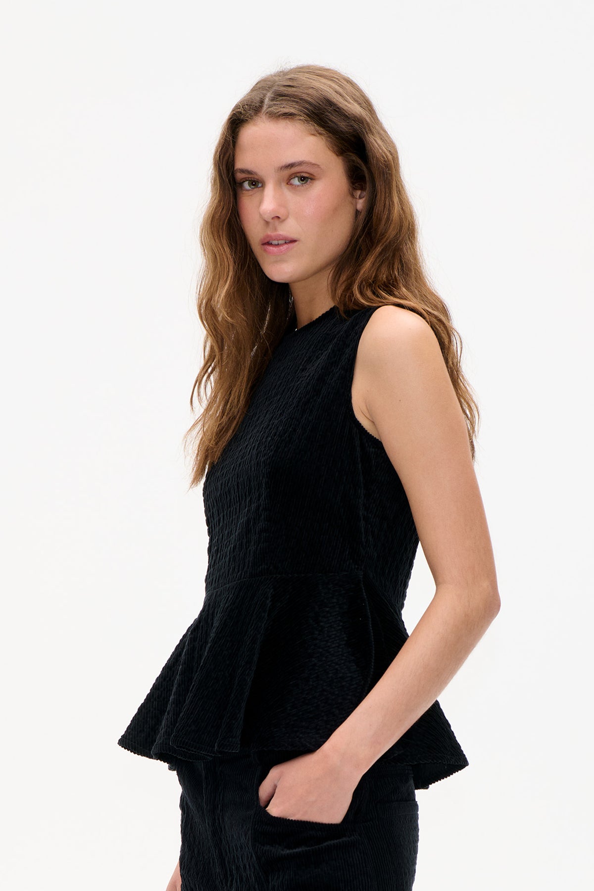 Studio portrait of a model wearing a black sleeveless textured top with a high neckline and flared peplum hem.