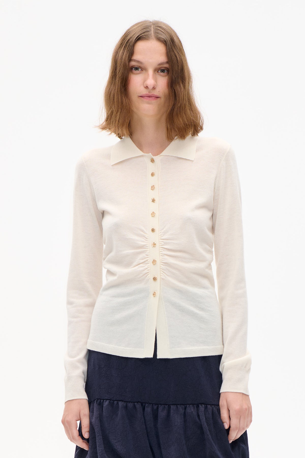 Front view of a cream long-sleeved knit cardigan with a pointed collar, decorative gold buttons and ruched sides, worn over a navy dress.