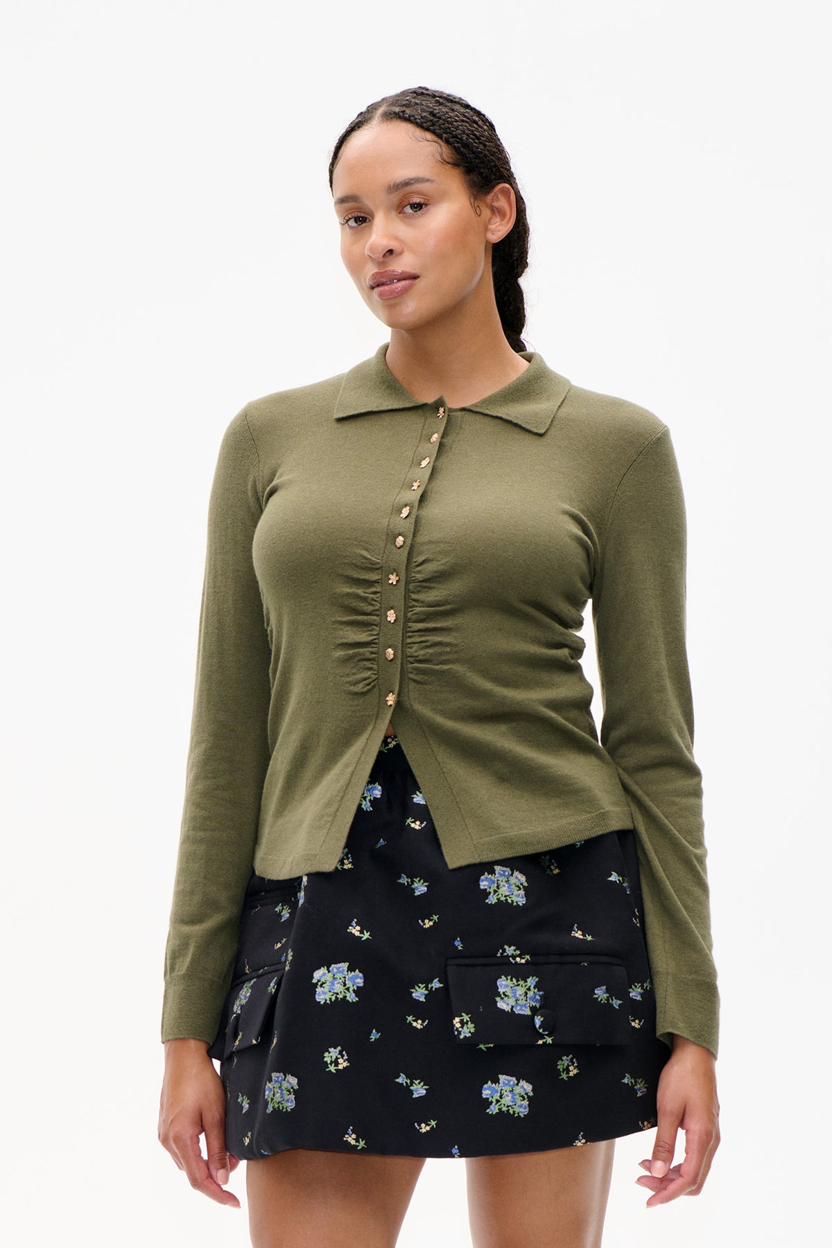 Front view of an olive-green collared cardigan with ruched detailing and flower-shaped buttons, worn over a black floral skirt.