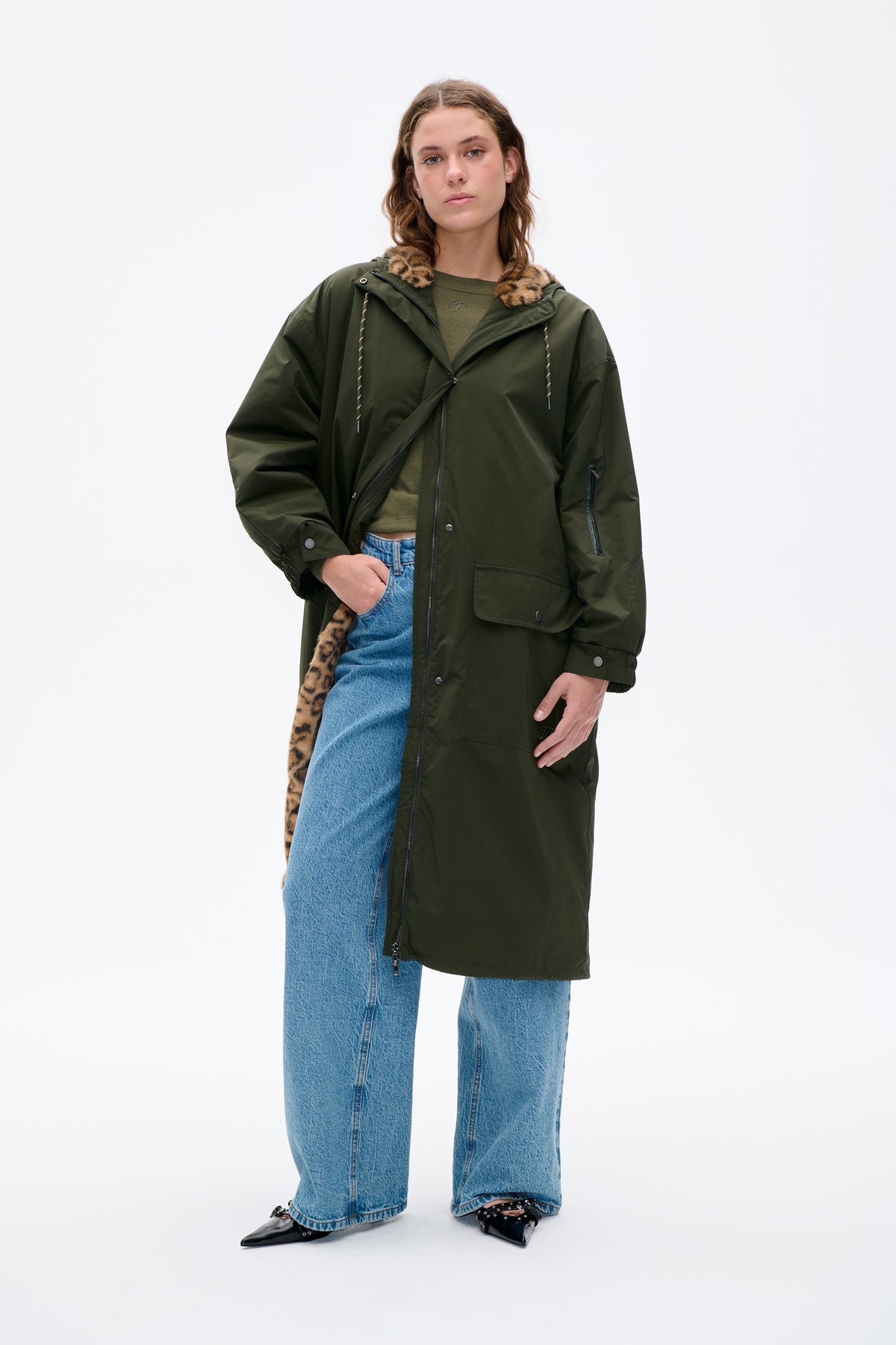 Front view of a model wearing a long dark olive coat with a leopard-print hood lining, layered over a green top and blue jeans.