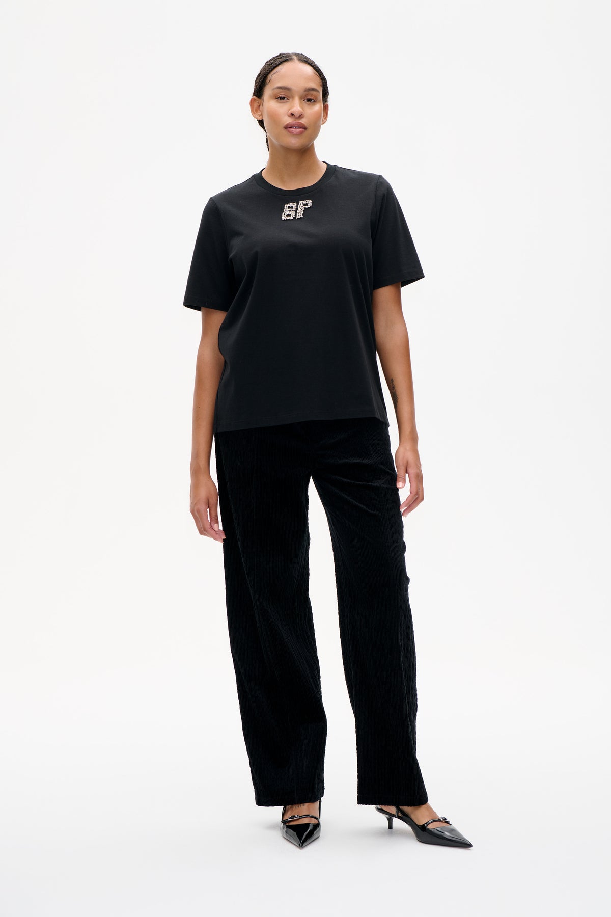 Vorderansicht eines schwarzen T-Shirts mit kurzen Ärmeln und Rundhalsausschnitt, verziert mit glitzernden BP-Initialen auf der Brust, kombiniert mit schwarzen Hosen mit weitem Bein.