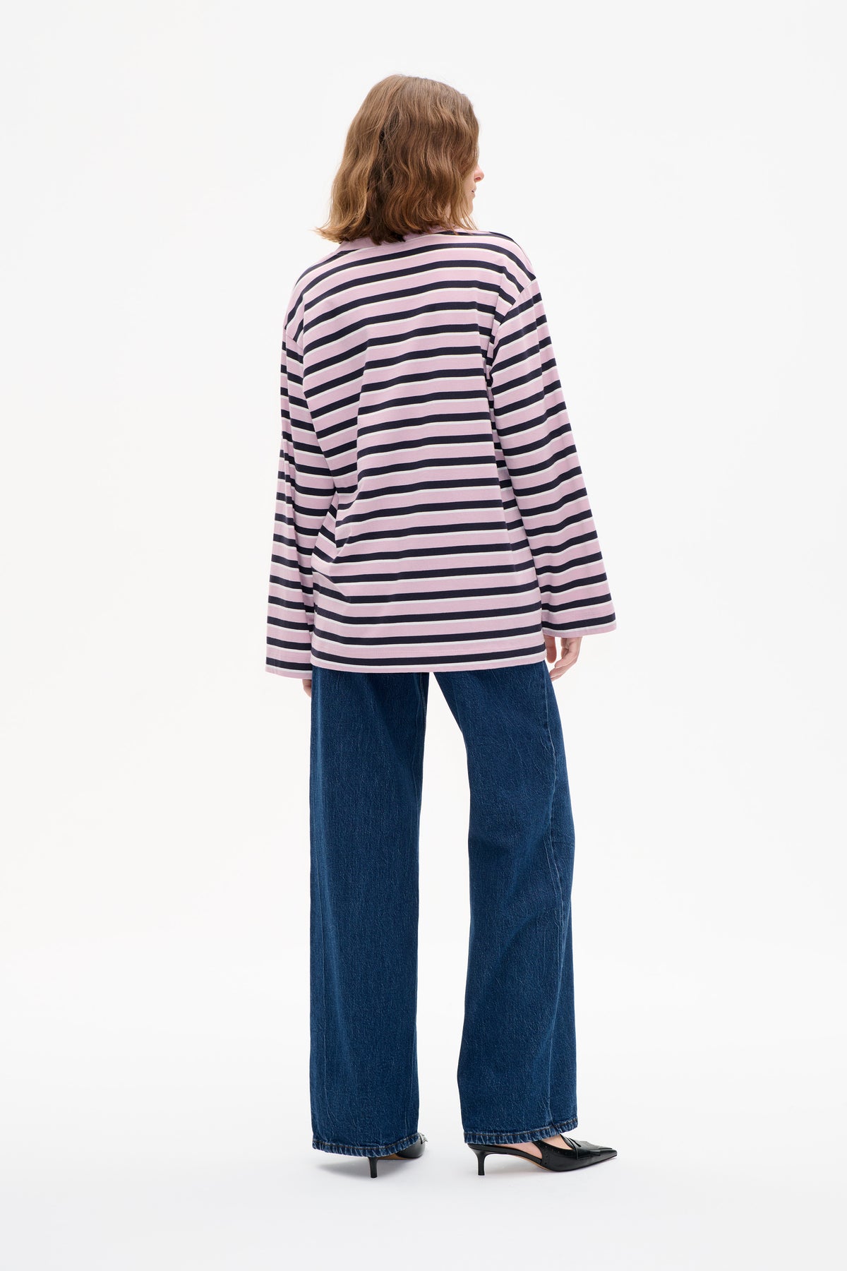 Model van achteren draagt een losse roze top met lange mouwen en marineblauwe, dunne crème horizontale strepen, gecombineerd met wijde blauwe jeans.