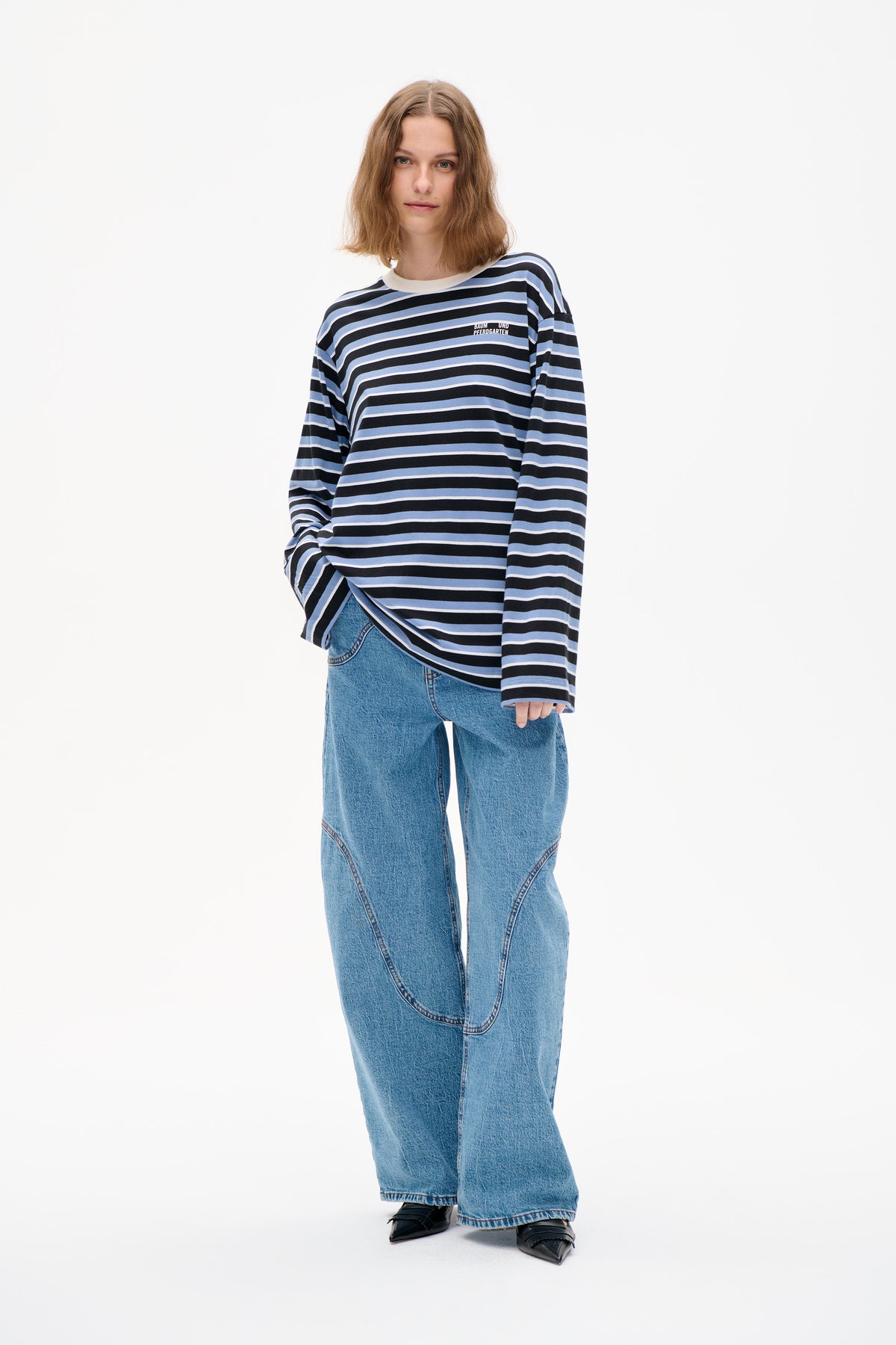 Front view of a model wearing a black, pale blue and white striped long-sleeve top with a small chest logo and wide-leg blue jeans.