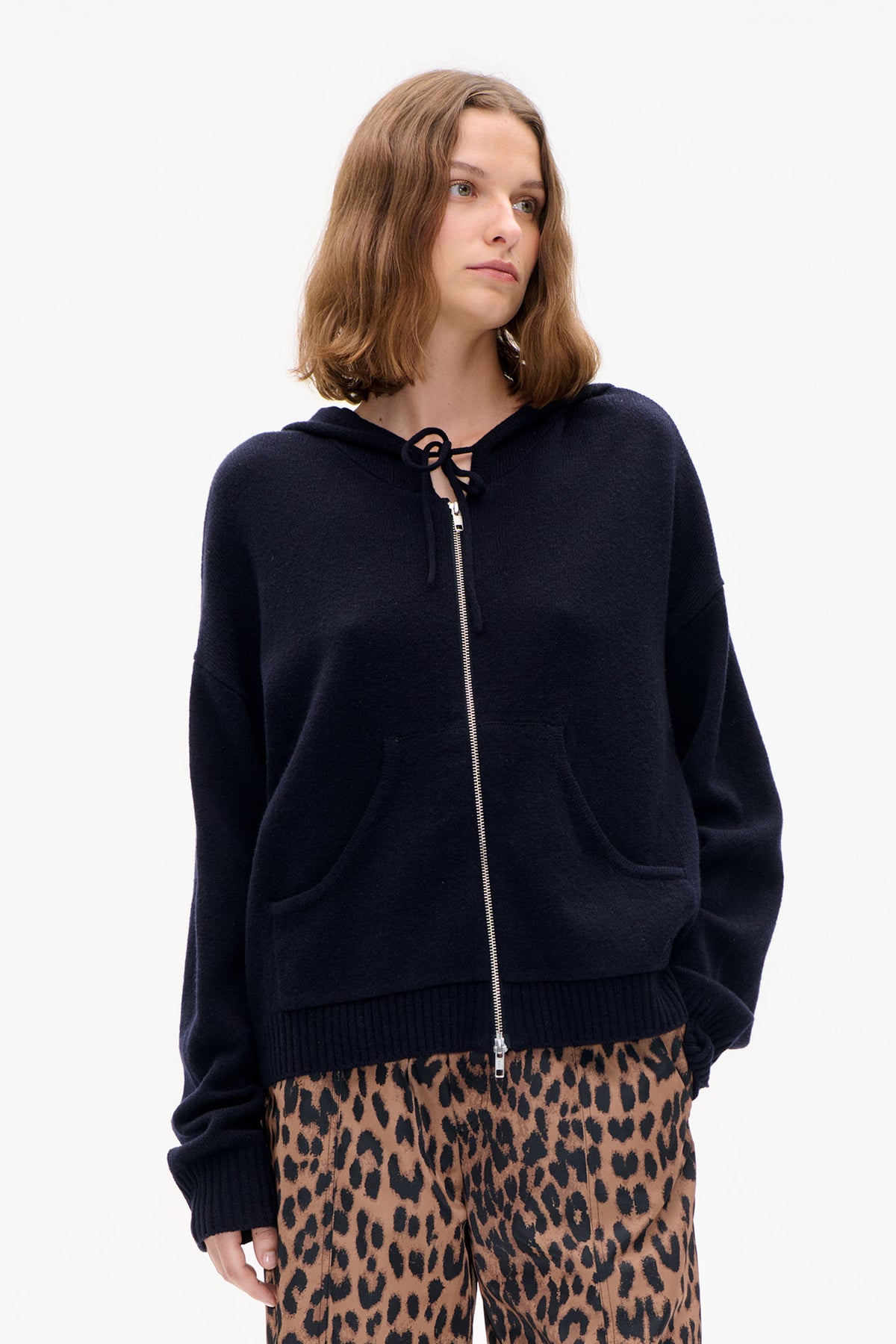 Close-up front view of a navy zip-up hooded knit jacket with a drawstring hood and large pockets, worn over leopard-print trousers.