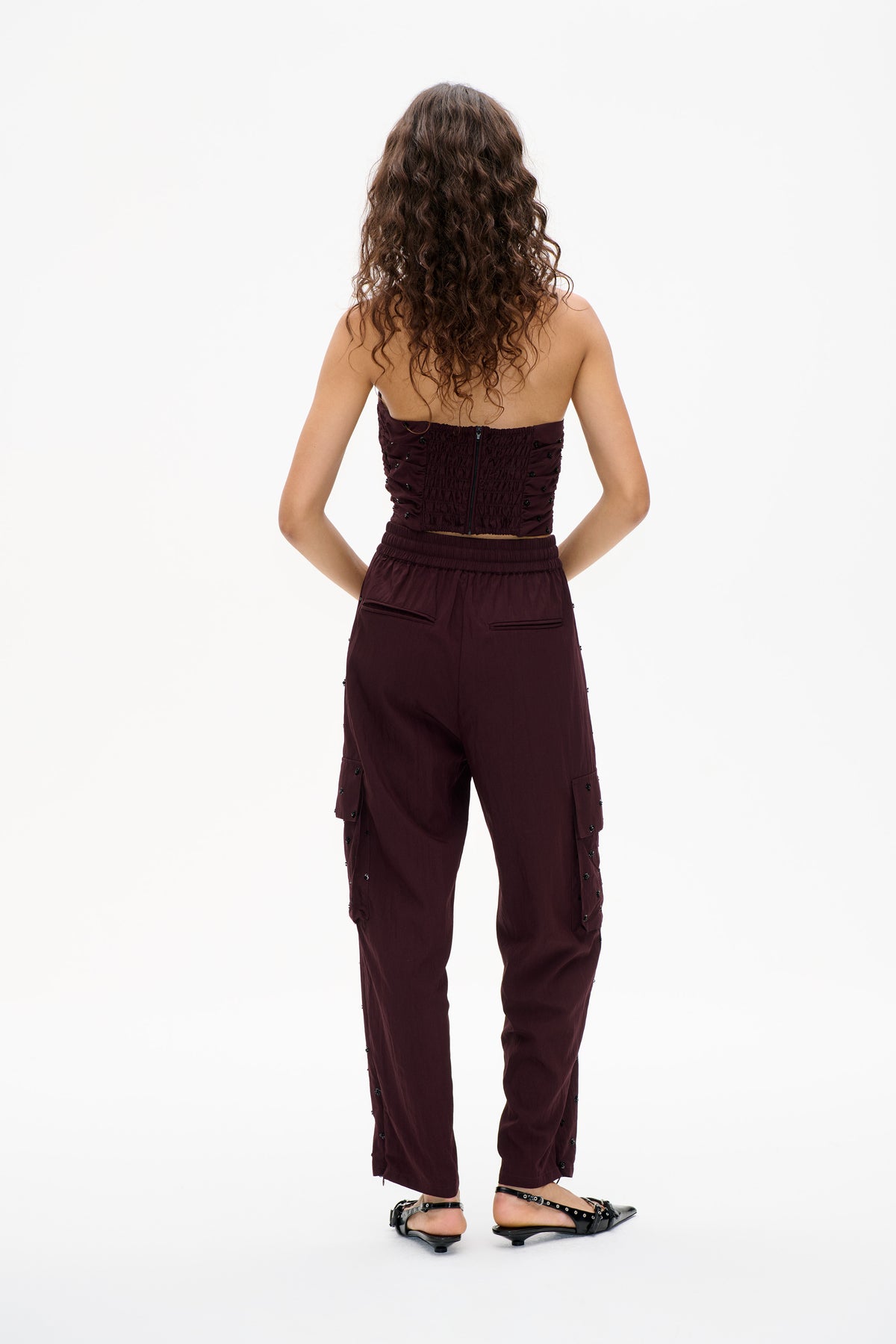 Achteraanzicht van een model in een bordeauxrode bandeau-top met smock en centrale ritssluiting, gecombineerd met een bijpassende wijde broek met zwarte details.