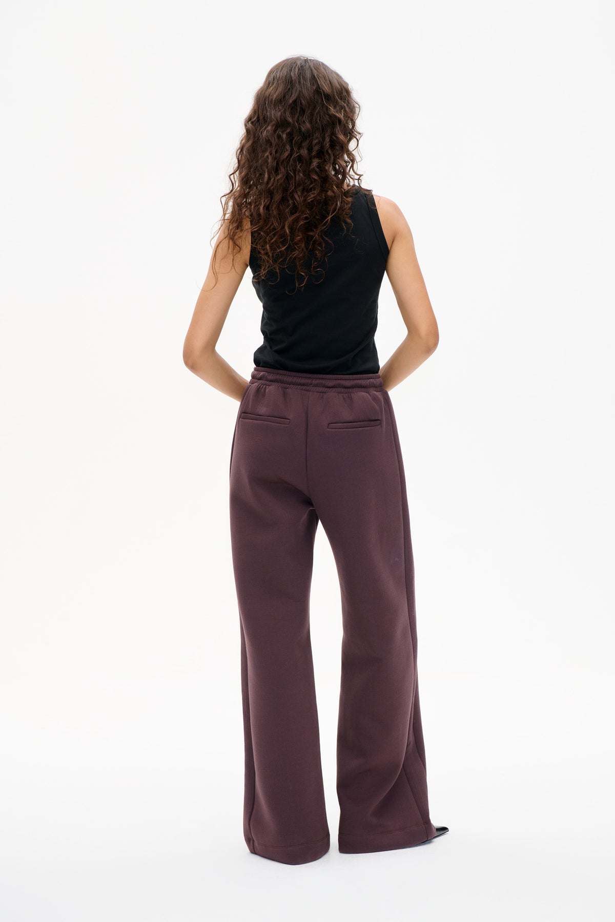 Achteraanzicht van een zwarte mouwloze top en een pruimkleurige broek met wijde pijpen, elastische tailleband en twee paspelzakken.
